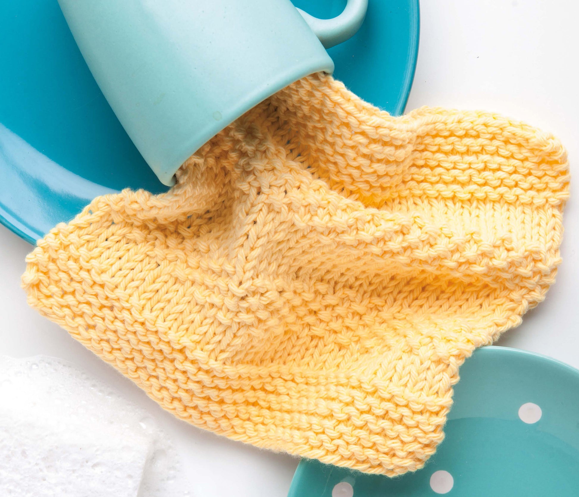 A yellow knitted dishcloth peeks from a light blue cup on blue plates atop a white background—perfect inspiration from Leisure Arts' "Cool Stuff Teach Me to Knit—13 Projects for Kids 9-14.