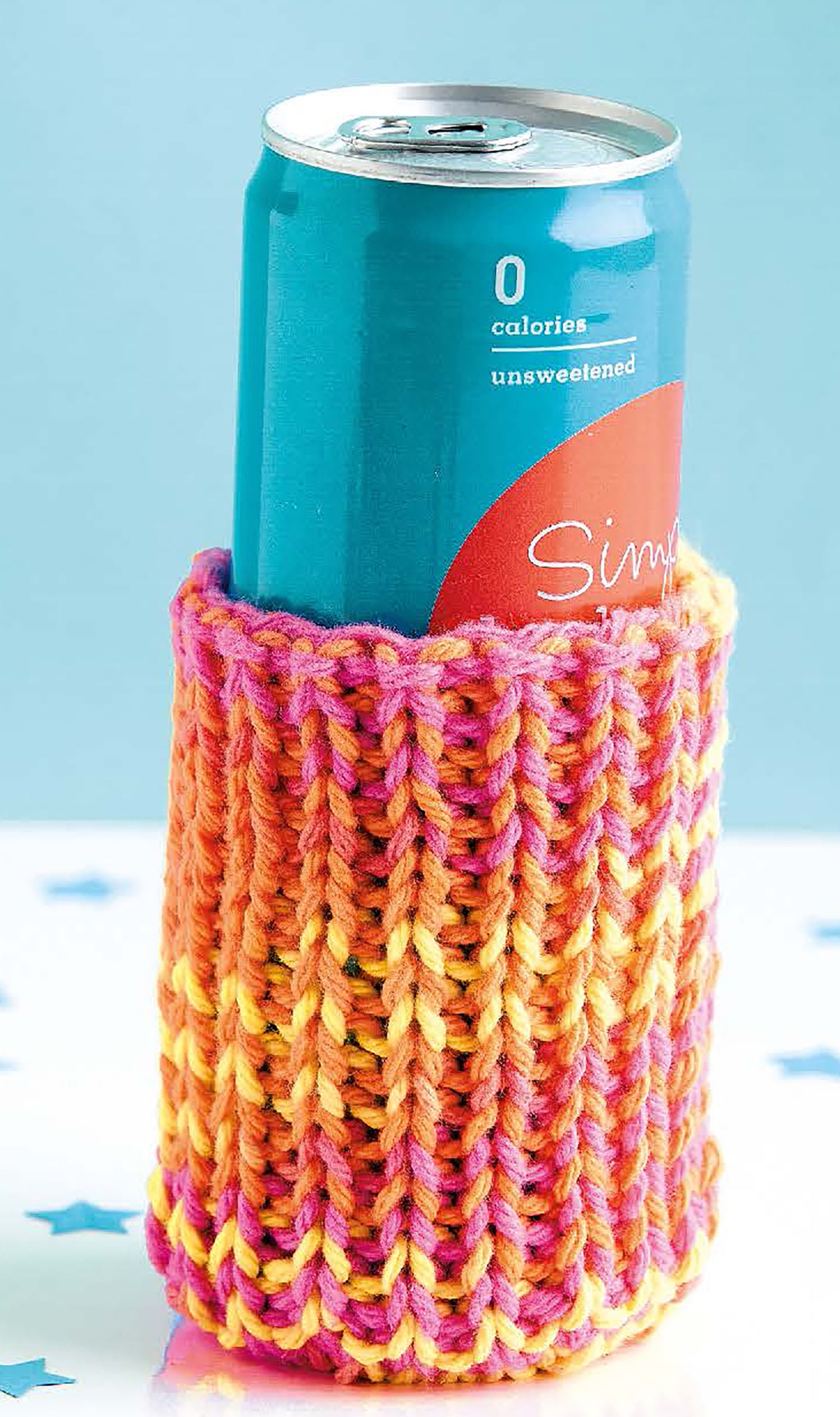 A teal soda can in a hand-knitted cozy of pink, yellow, and orange yarn sits on a blue background with star confetti. Try this fun beginner project from Leisure Arts’ "Cool Stuff Teach Me to Knit—13 Projects for Kids 9-14.