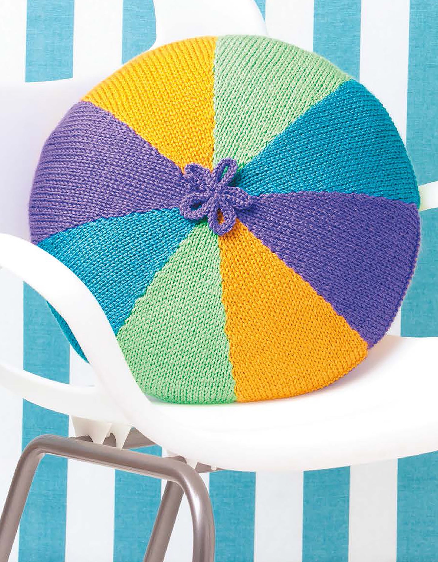 A round, knitted cushion with colorful segments and a purple flower detail from Leisure Arts' "Cool Stuff Teach Me to Knit—13 Projects for Kids 9-14" rests on a white chair against a blue and white striped background.