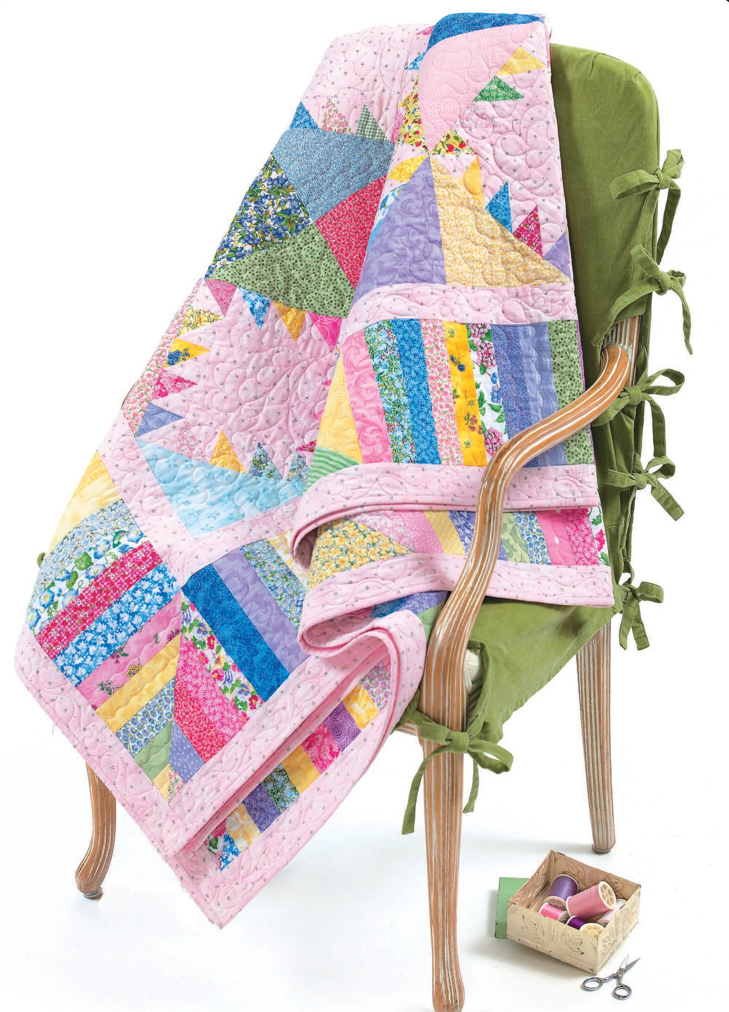 A Me & My Sister's Fun Quilts by Leisure Arts, featuring pastel geometric and floral designs inspired by beginner patterns, is draped over a green armchair; nearby sits a small sewing supplies box.