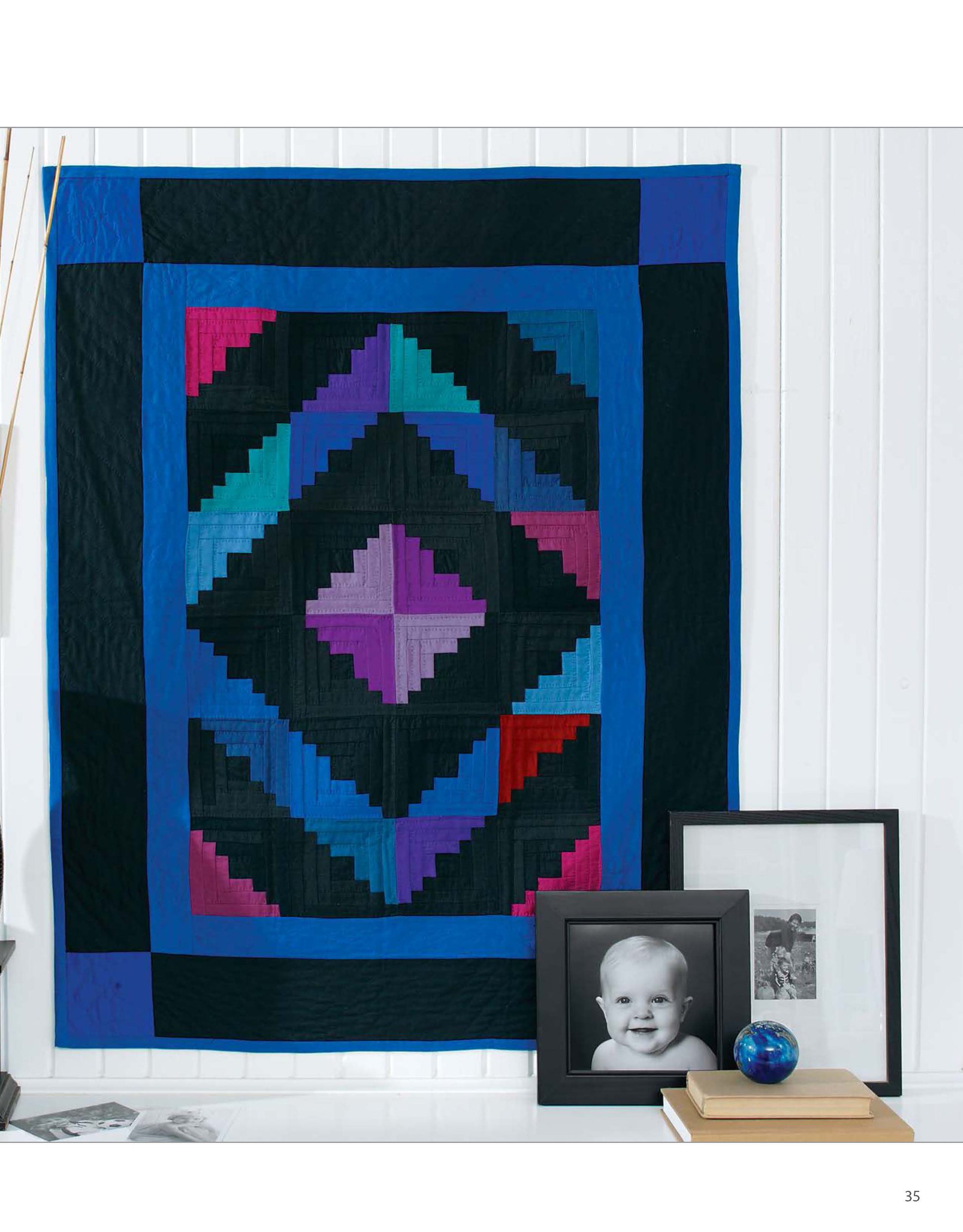 The Classic Quilts by Leisure Arts, featuring modern blue, purple, black, and red geometric designs, hangs on a white wall above a shelf with framed black-and-white photos and a small blue glass sphere.