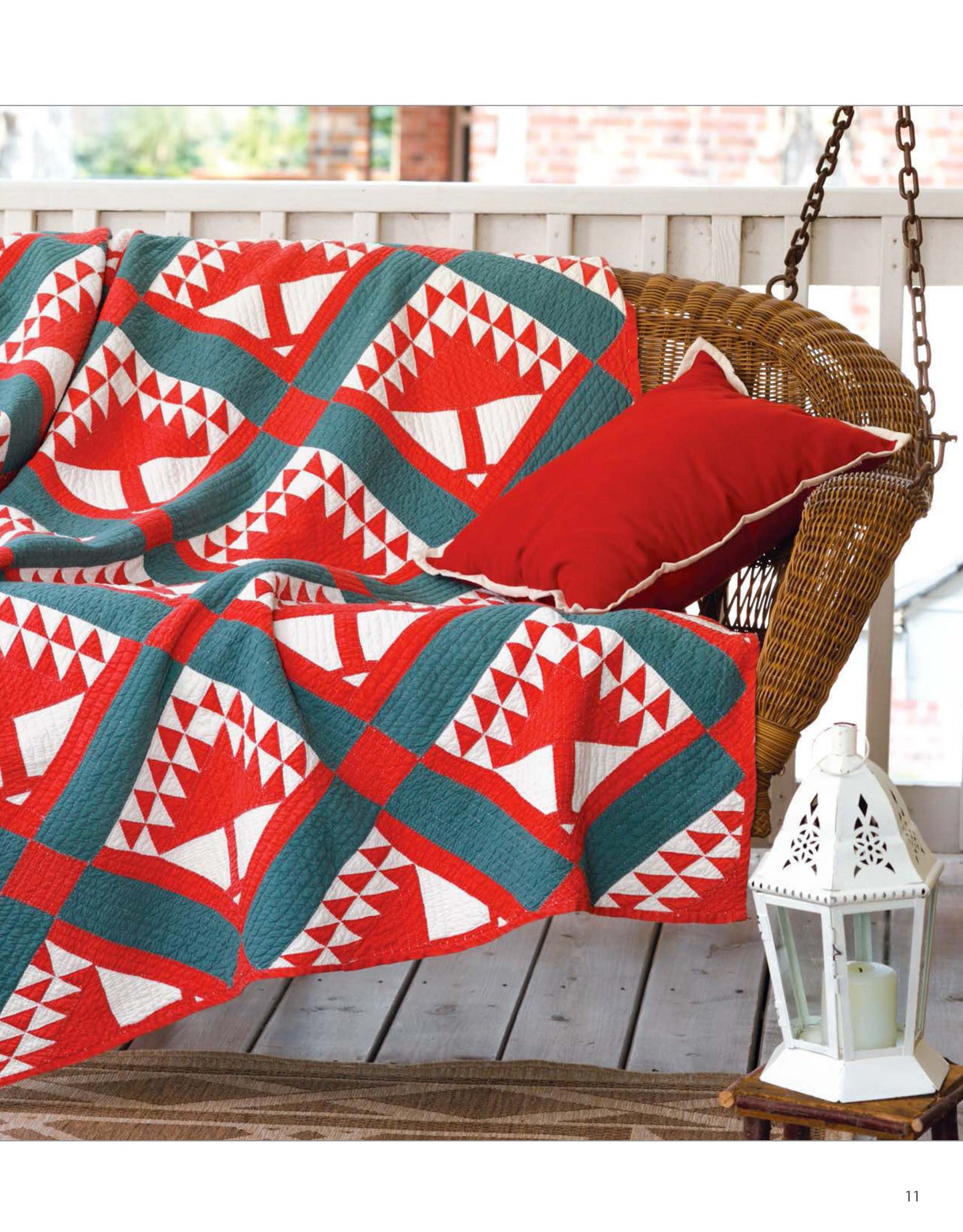 A wicker porch swing with a Leisure Arts Classic Quilts geometric-patterned quilt in red, white, and teal draped over it, a red cushion, and a white decorative lantern on the nearby wooden floor.