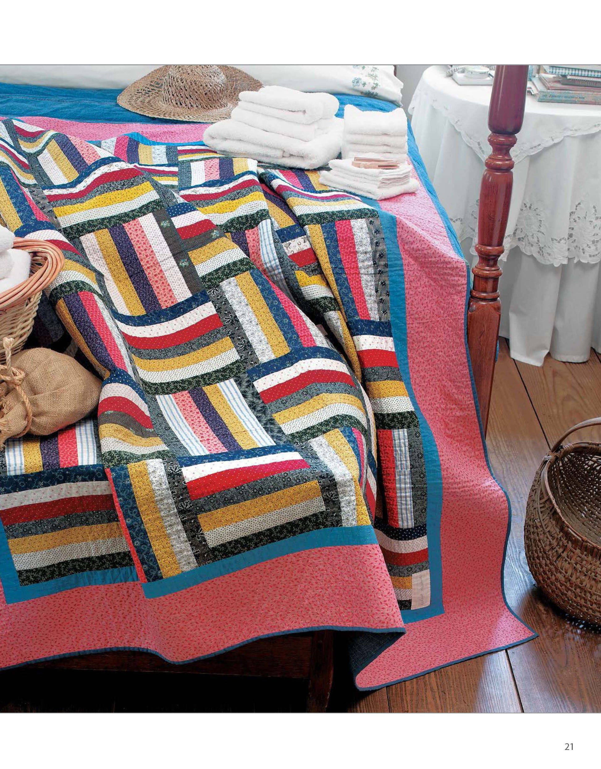 A Classic Quilts coverlet by Leisure Arts with striped patchwork design covers a wooden bed. Towels, baskets, and a straw hat rest nearby. The room includes a wooden floor and a white table draped with a lacy cloth.