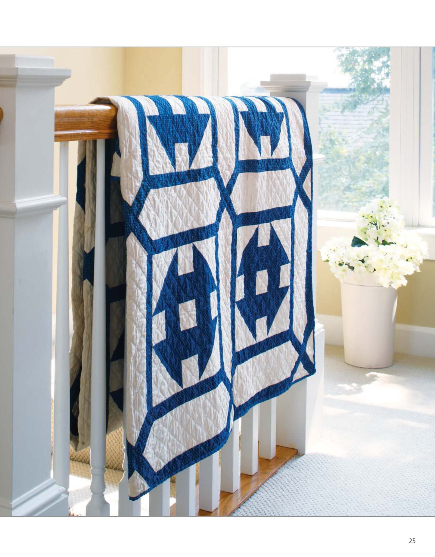 The Classic Quilts by Leisure Arts, featuring blue and white geometric patterns, is draped over a wooden staircase railing in a bright room with a window and a vase of white flowers.