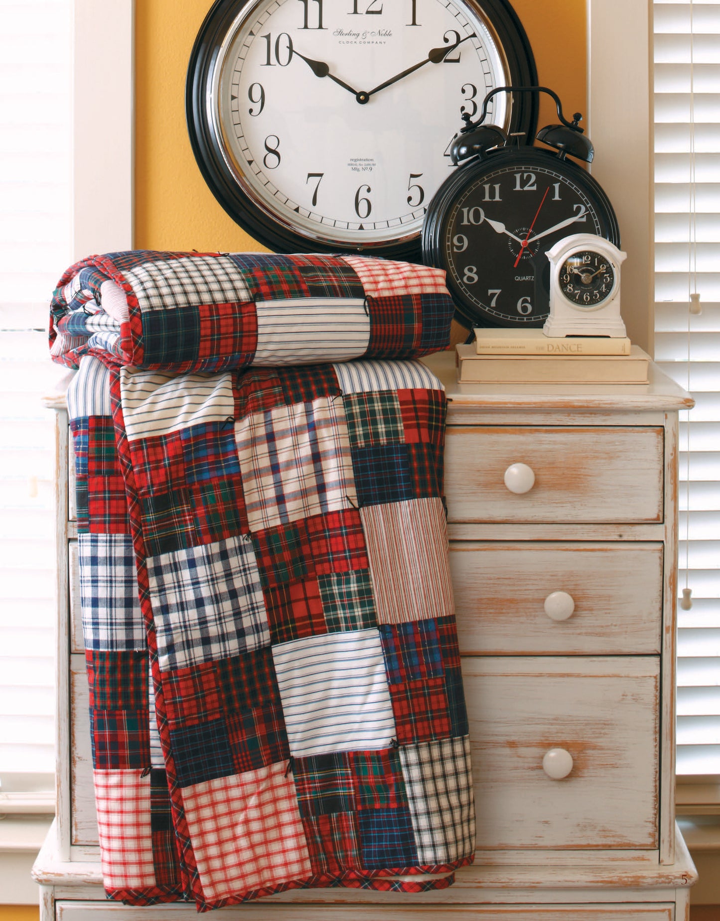 The Easy Timeless Quilts Digital Download by Leisure Arts is displayed on a distressed white dresser with two black-and-white clocks and a small silver clock on top, against closed blinds and a yellow wall in the background.