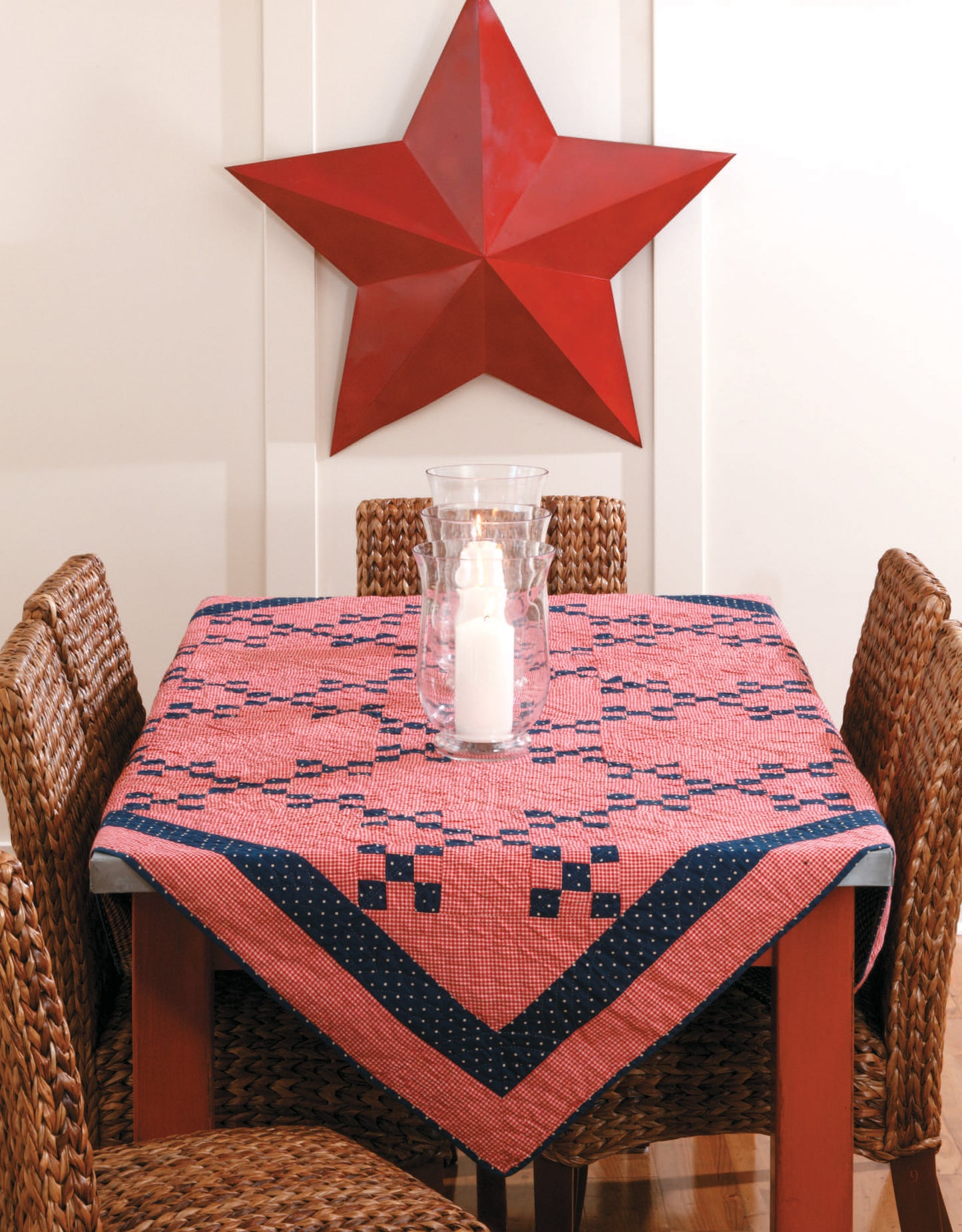A square table features a quilted tablecloth made with patterns from the Easy Timeless Quilts Digital Download by Leisure Arts. Four wicker chairs encircle it, with a glass hurricane candle holder at the center and a large red star on the wall.