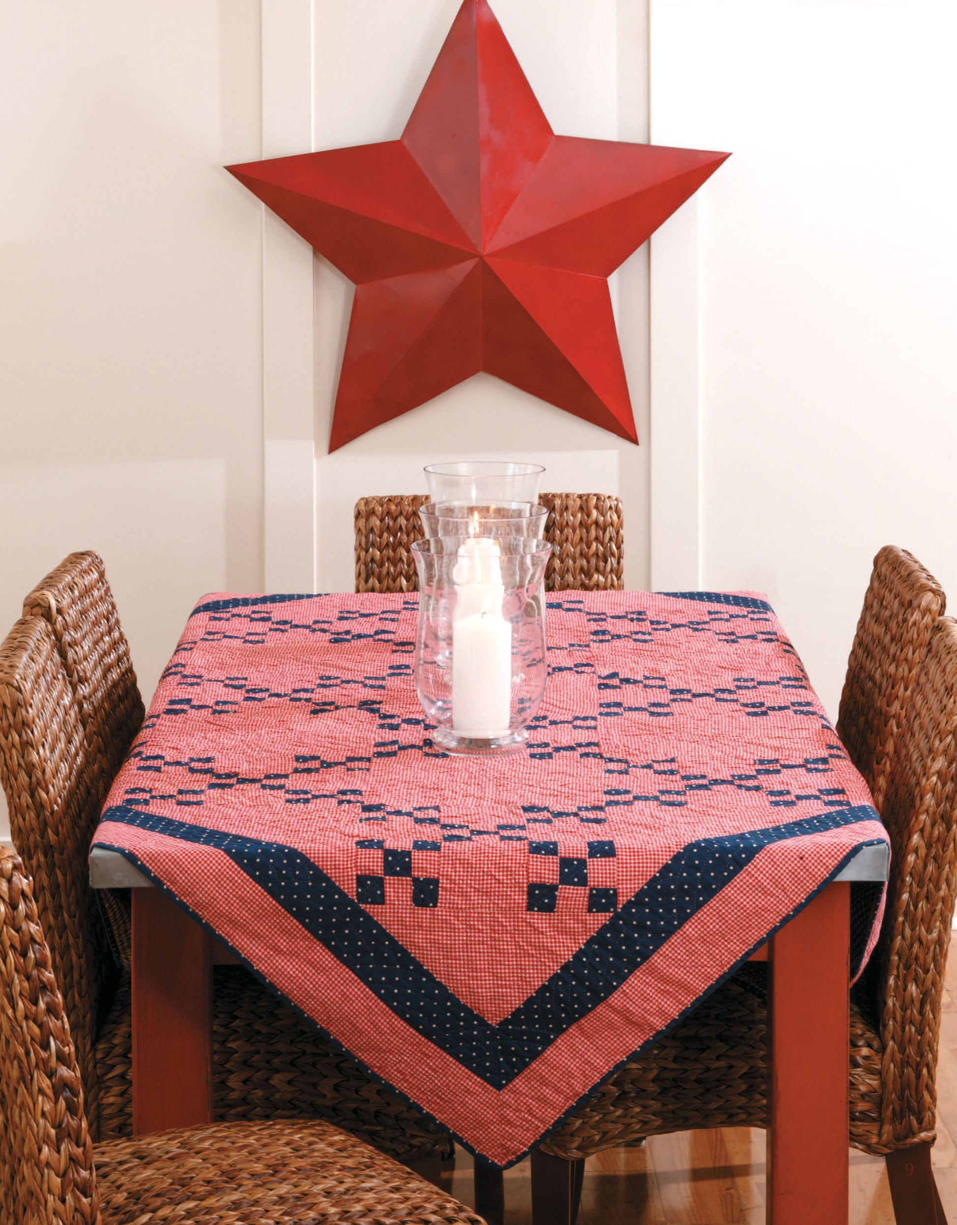 A square table features a quilted tablecloth made with patterns from the Easy Timeless Quilts Digital Download by Leisure Arts. Four wicker chairs encircle it, with a glass hurricane candle holder at the center and a large red star on the wall.