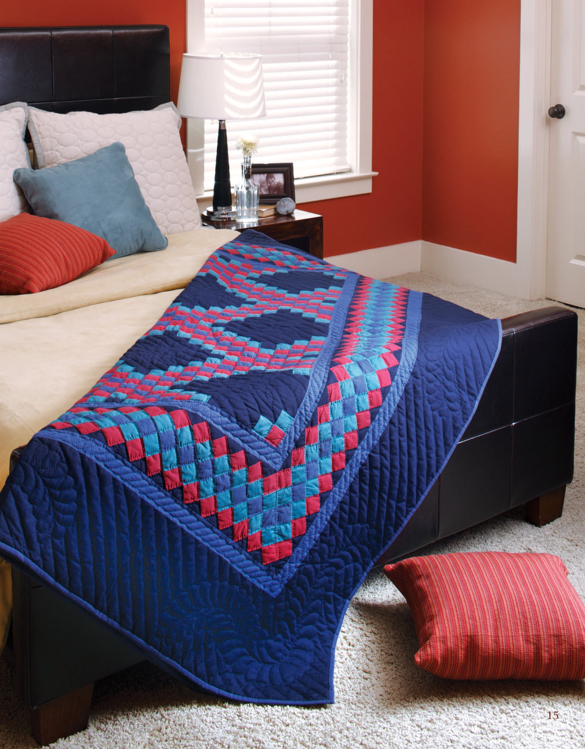 A cozy bedroom features neutral bedding topped with colorful pillows. The bed showcases a vibrant blue, red, and aqua quilt made from Leisure Arts’ Easy Timeless Quilts Digital Download, with a matching pillow on the floor.