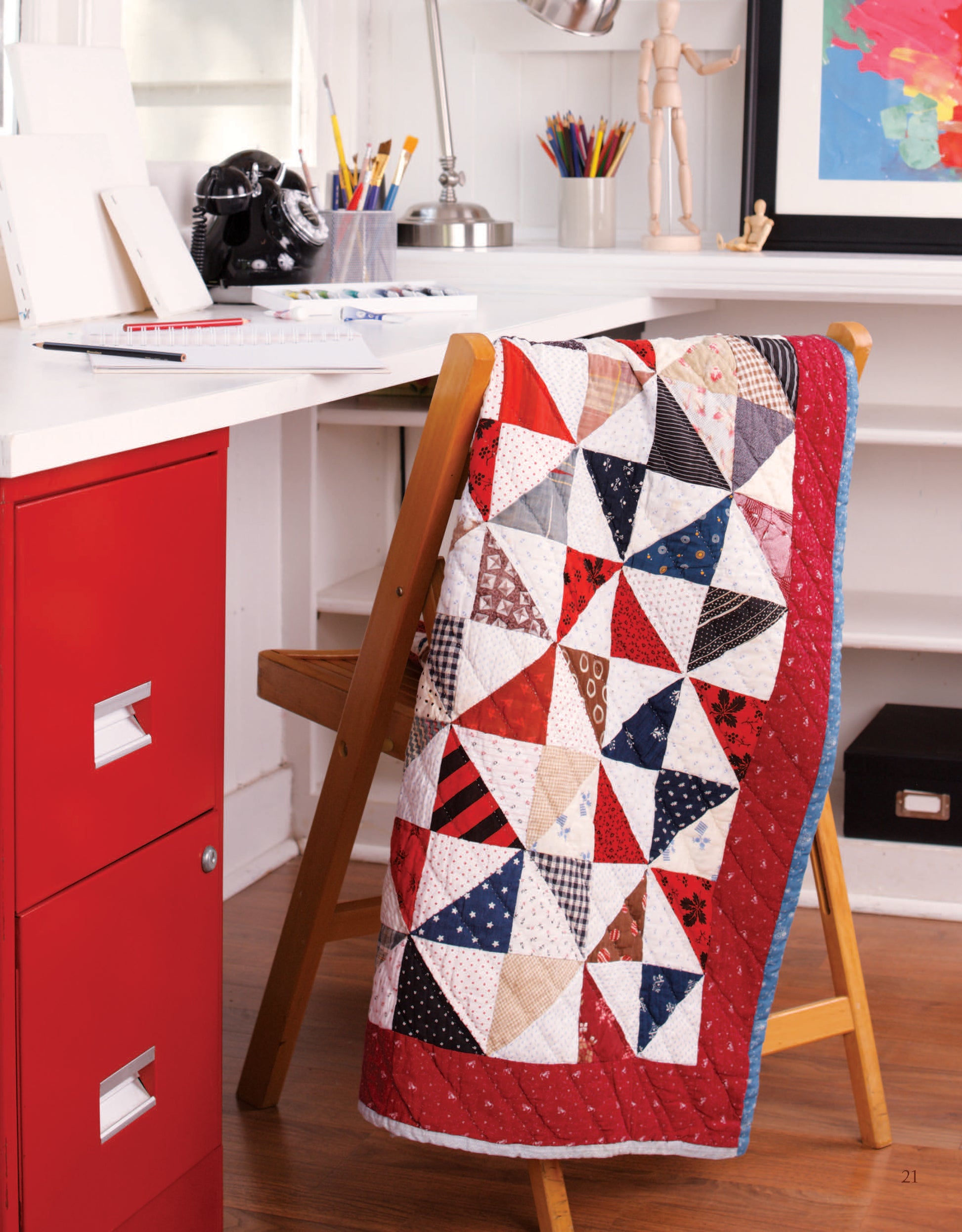 A quilt made with the Easy Timeless Quilts Digital Download by Leisure Arts, featuring red, white, and blue triangles, drapes over a chair next to a red filing cabinet in a bright workspace filled with art supplies and vibrant decor.