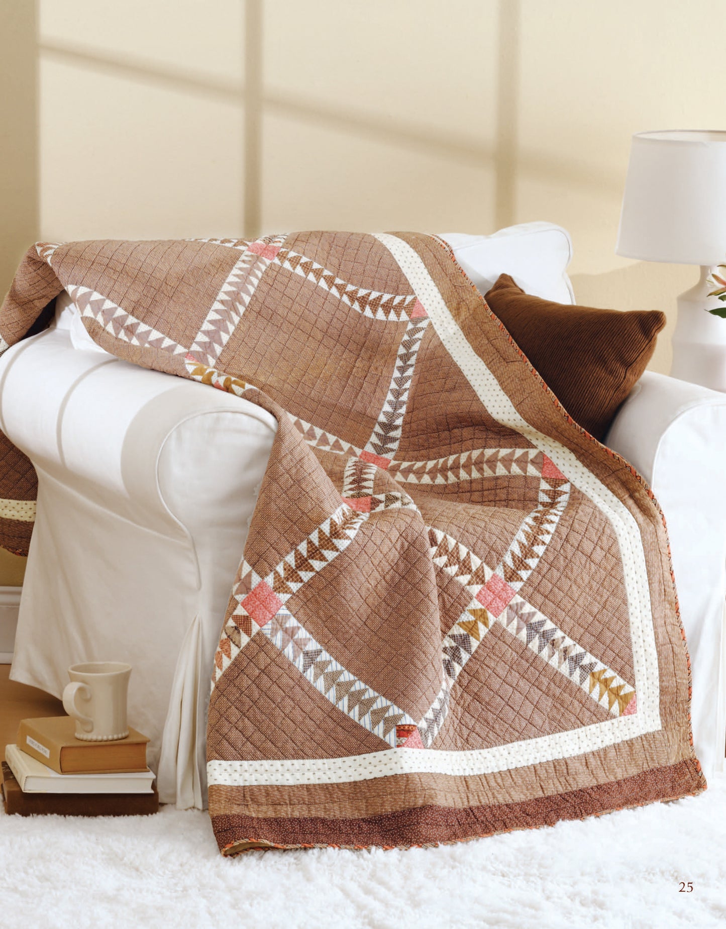 The Easy Timeless Quilts Digital Download by Leisure Arts, featuring brown geometric patterns, is displayed over a white armchair beside a brown cushion, lamp, and side table with a book and mug.