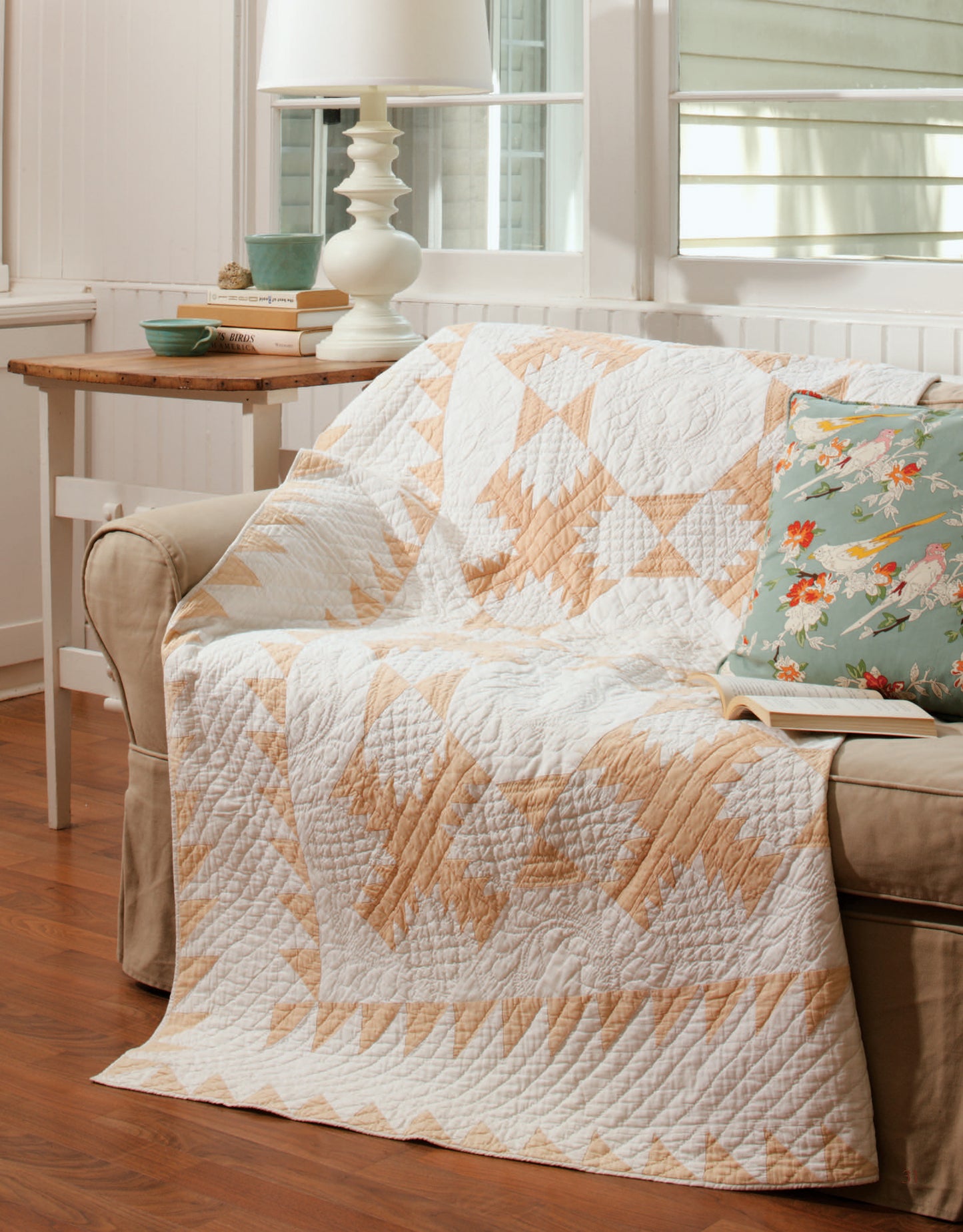 The Easy Timeless Quilts Digital Download by Leisure Arts is shown as a modern beige and white patchwork quilt draped over a sofa with a floral pillow, beside a wooden side table with a lamp in a cozy, sunlit living room.