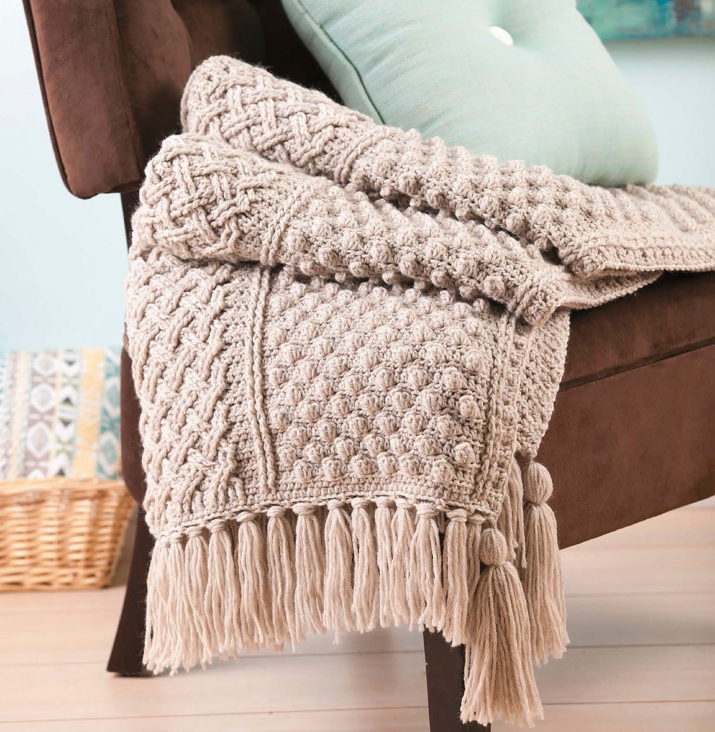 The brown chair features a cozy throw from the "Afghans with Style Digital Download" by Leisure Arts, paired with a light blue cushion, while a woven basket sits in the softly lit background.