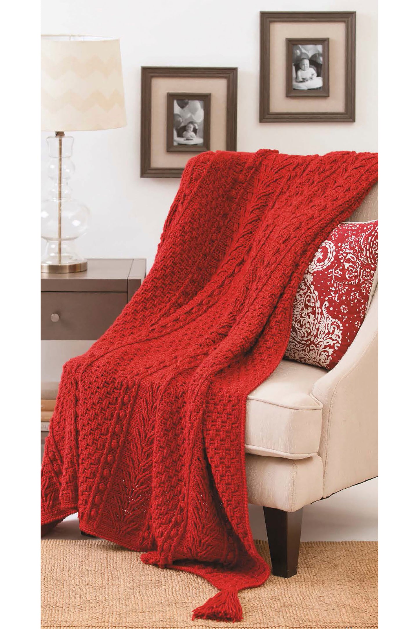 A cozy red knit blanket from Leisure Arts' Afghans with Style Digital Download drapes over a cream armchair with a red and white pillow. A lamp and framed black-and-white photos are visible on the wall and side table in the background.