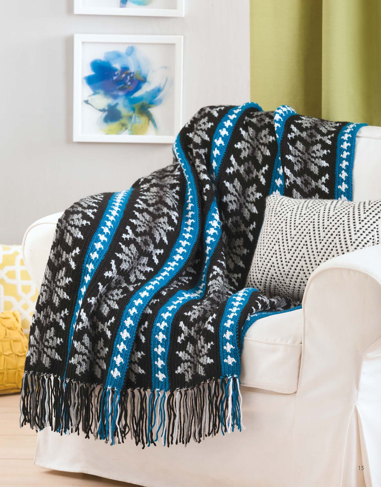 A stylish crocheted throw from the "Afghans with Style Digital Download" by Leisure Arts, featuring black, gray, white, and blue stripes with fringed edges, is draped over a white chair. Floral art and a yellow pillow add touches of color to the room.