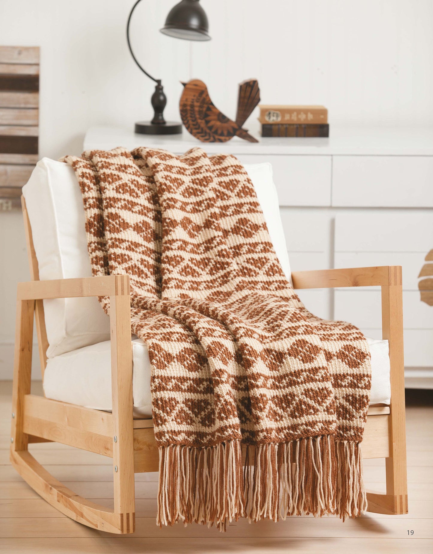 A cozy brown and cream Aran crochet throw from Leisure Arts' "Afghans with Style" digital download drapes over a light wooden rocking chair with white cushions; a white dresser, lamp, and decorative bird are in the background.