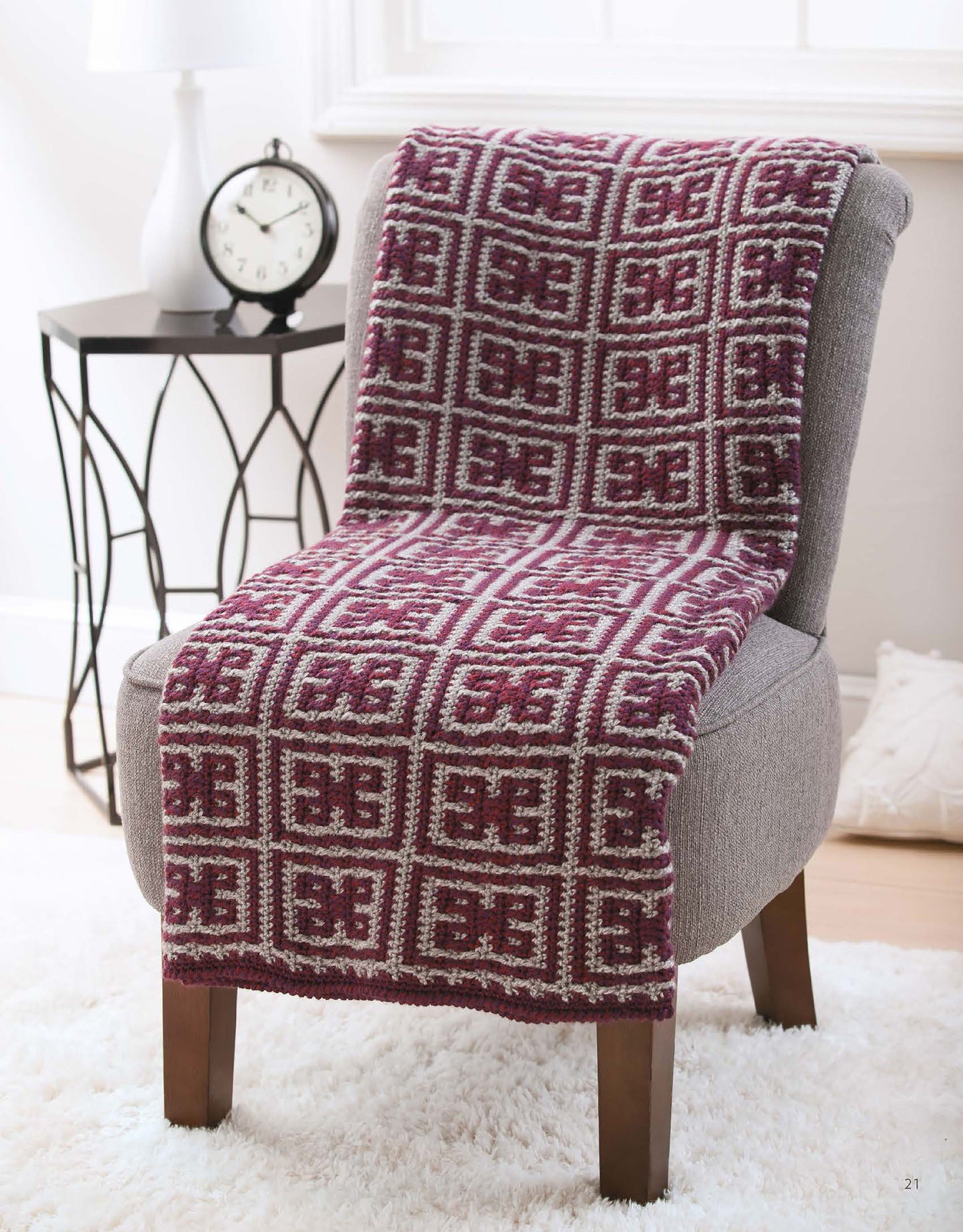 A gray chair draped with a maroon and white Fair Isle blanket from Afghans with Style by Leisure Arts sits beside a side table with a white clock and vase in a bright, softly lit room.