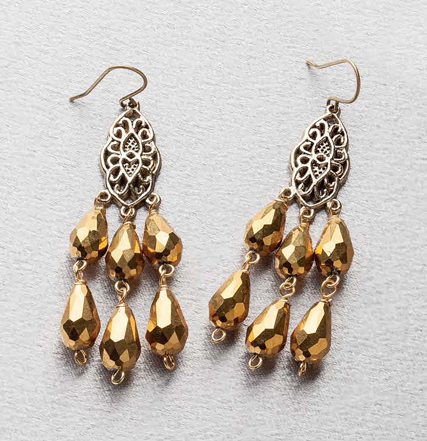 Jewelry for the Stars by Leisure Arts: Elegant gold dangle earrings with ornate filigree tops and three rows of teardrop-shaped beads, perfect for pairing with a statement necklace, displayed on a light gray surface.