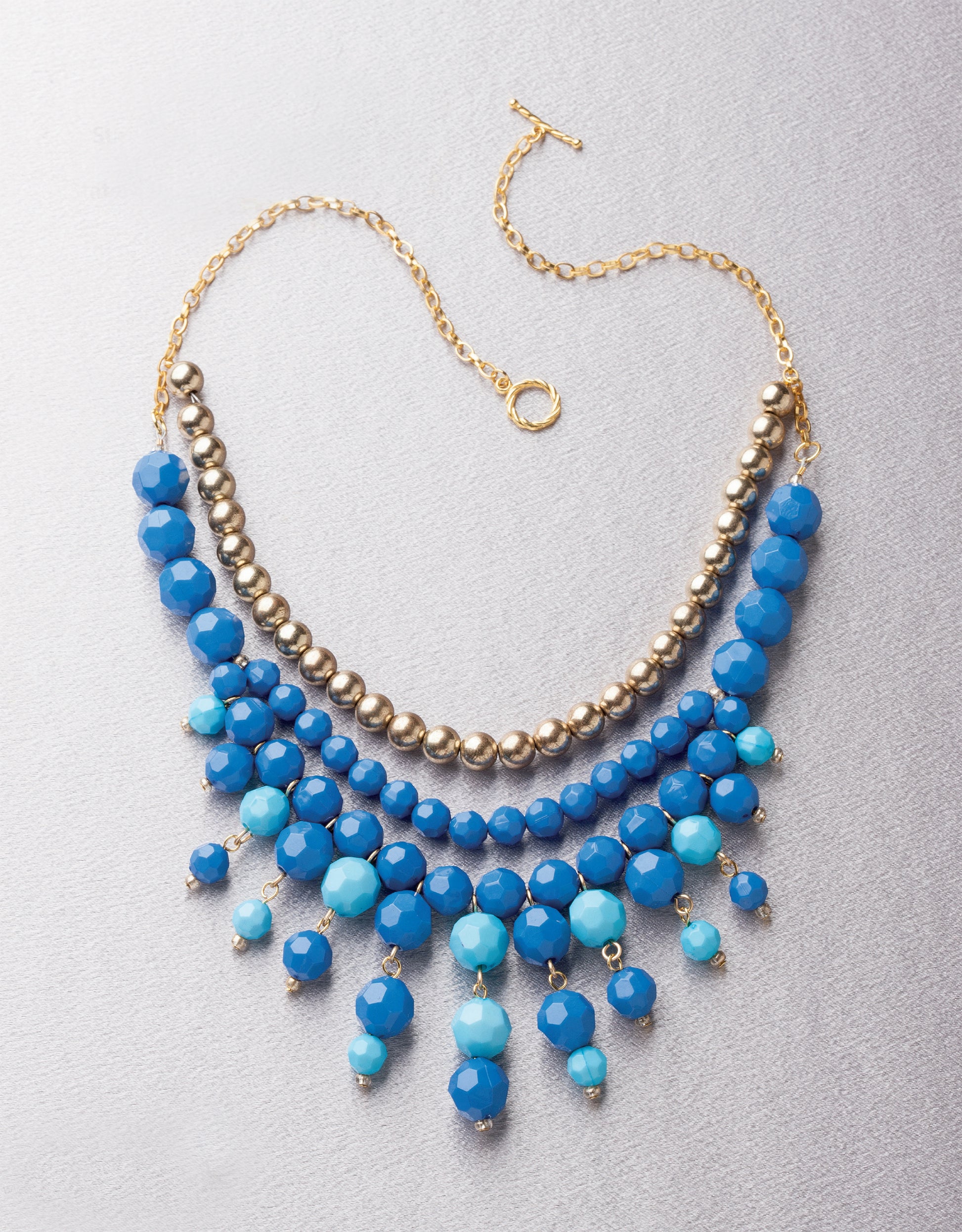 The Jewelry for the Stars necklace by Leisure Arts features three rows: metallic gold beads on top, two-tone blue beads in the middle, and dangling blue beads at the bottom. It is displayed flat on a light surface.