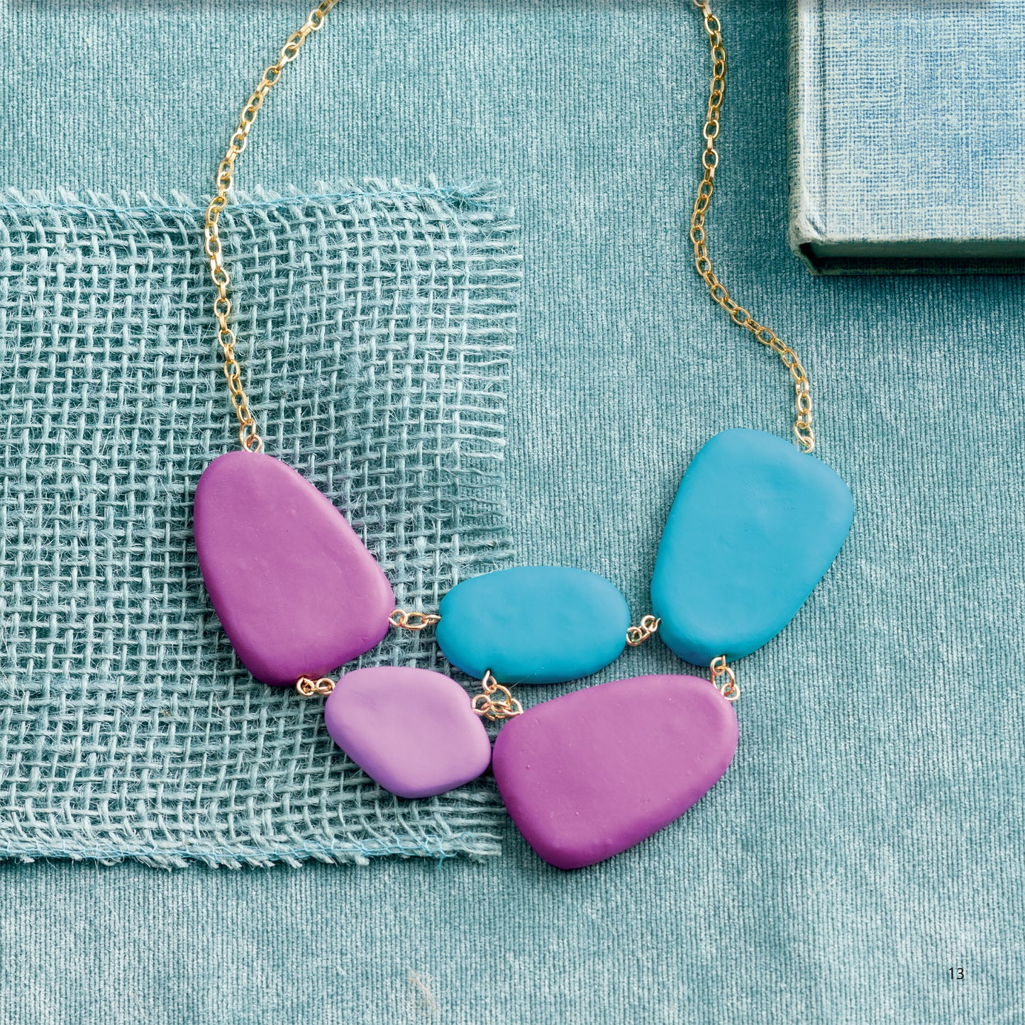 Bold Unique Jewelry by Leisure Arts features large, smooth purple and blue beads on a delicate gold chain, displayed on light blue textured fabric with a book and woven blue fabric in the background—a perfect statement piece for accessory lovers.