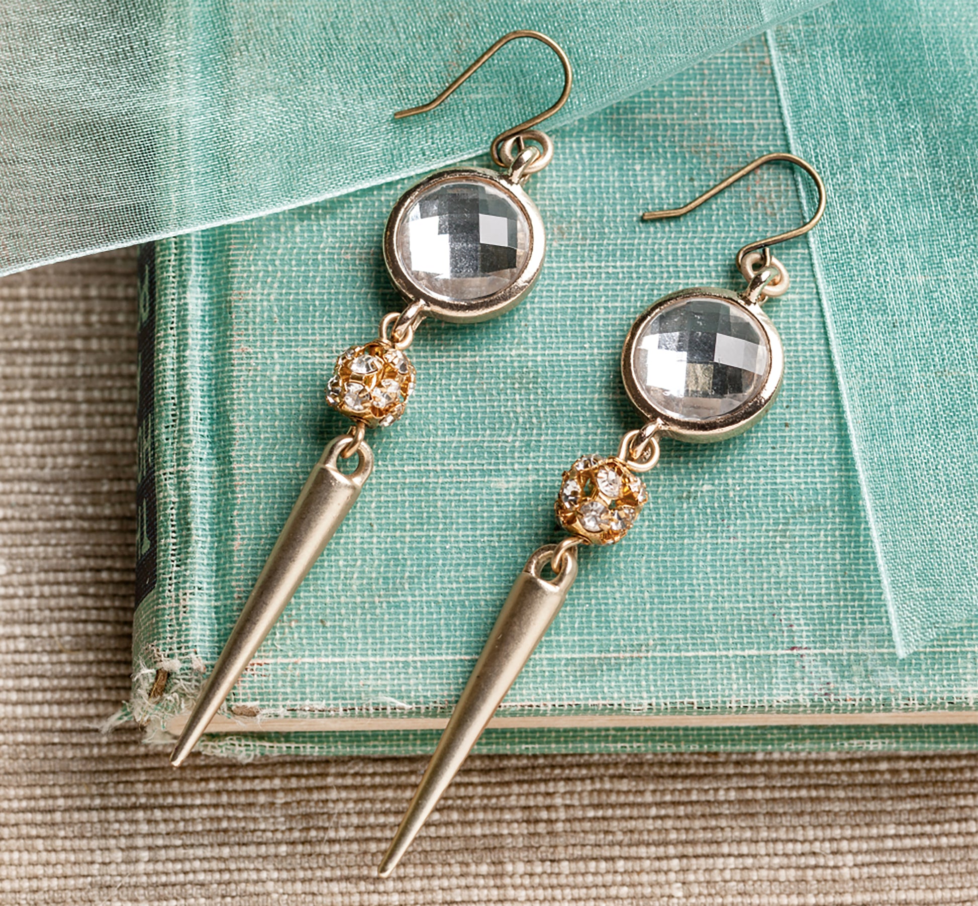 Leisure Arts' Bold Unique Jewelry features dangling earrings with faceted clear stones, gold-toned spikes, and crystal accents—eye-catching accessories stylishly displayed on stacked green books with sheer fabric.