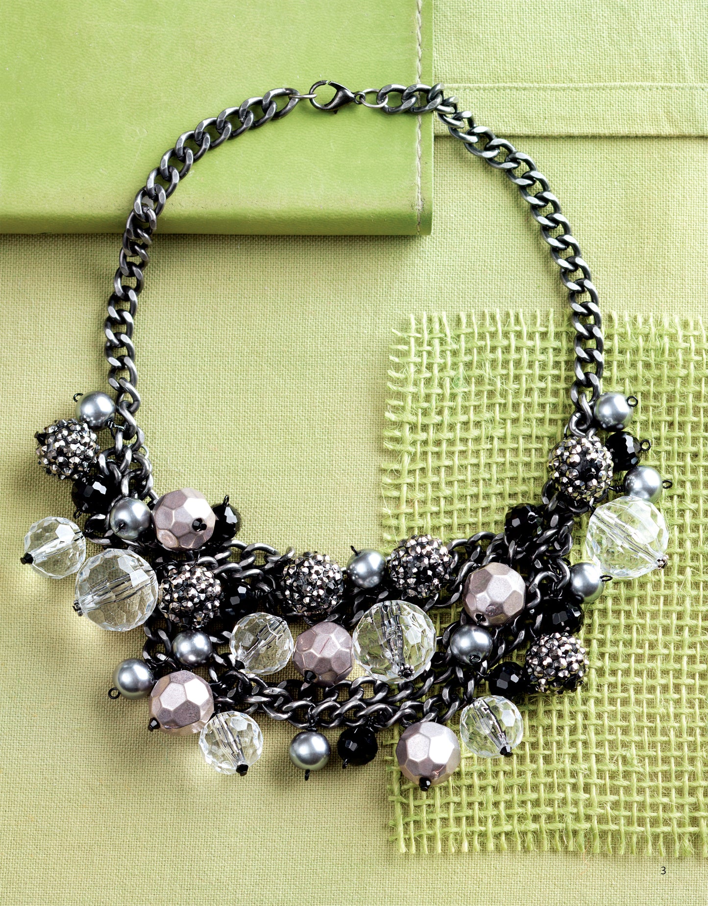 Bold Unique Jewelry by Leisure Arts—a chunky statement necklace with large clear, black, and metallic beads on a thick dark chain—is displayed on green fabric next to a light green book and woven mat.