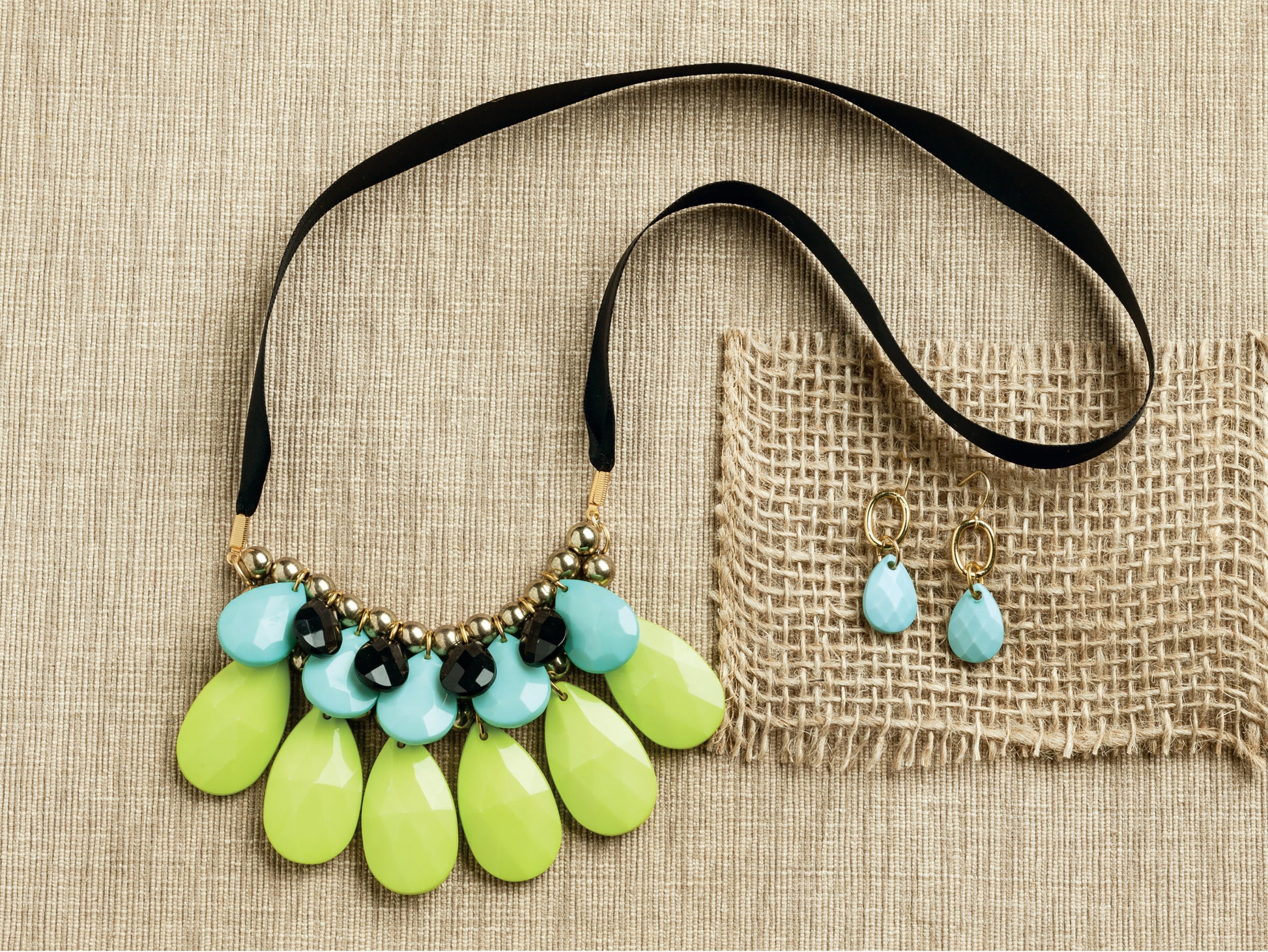 Bold Unique Jewelry by Leisure Arts features a statement necklace with blue and yellow-green teardrop beads and matching blue drop earrings, displayed on textured beige fabric and burlap—perfect for beginner jewelry lovers.