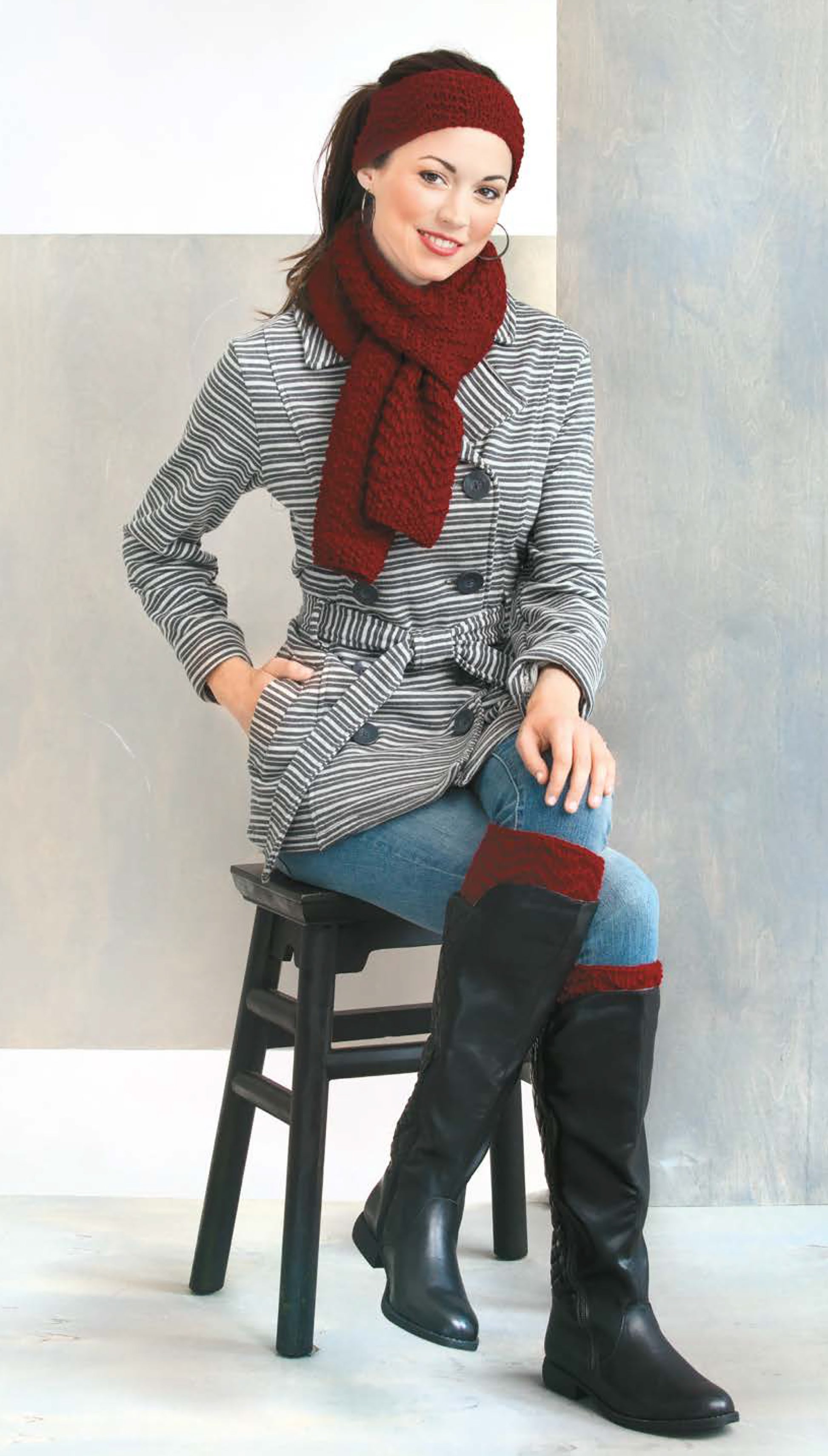 A woman models red knit boot cuffs, scarf, and ear warmers from the Cozy Fashion Accents Digital Download by Leisure Arts, paired with a striped coat and black boots, as she smiles at the camera against a neutral background.