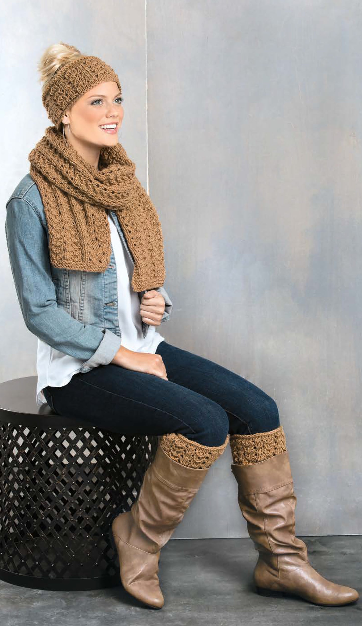 A woman models items from the Cozy Fashion Accents Digital Download by Leisure Arts, including a crochet scarf and ear warmers, paired with a denim jacket, white blouse, dark jeans, tan boots, and boot cuffs. She smiles, looking to the left.