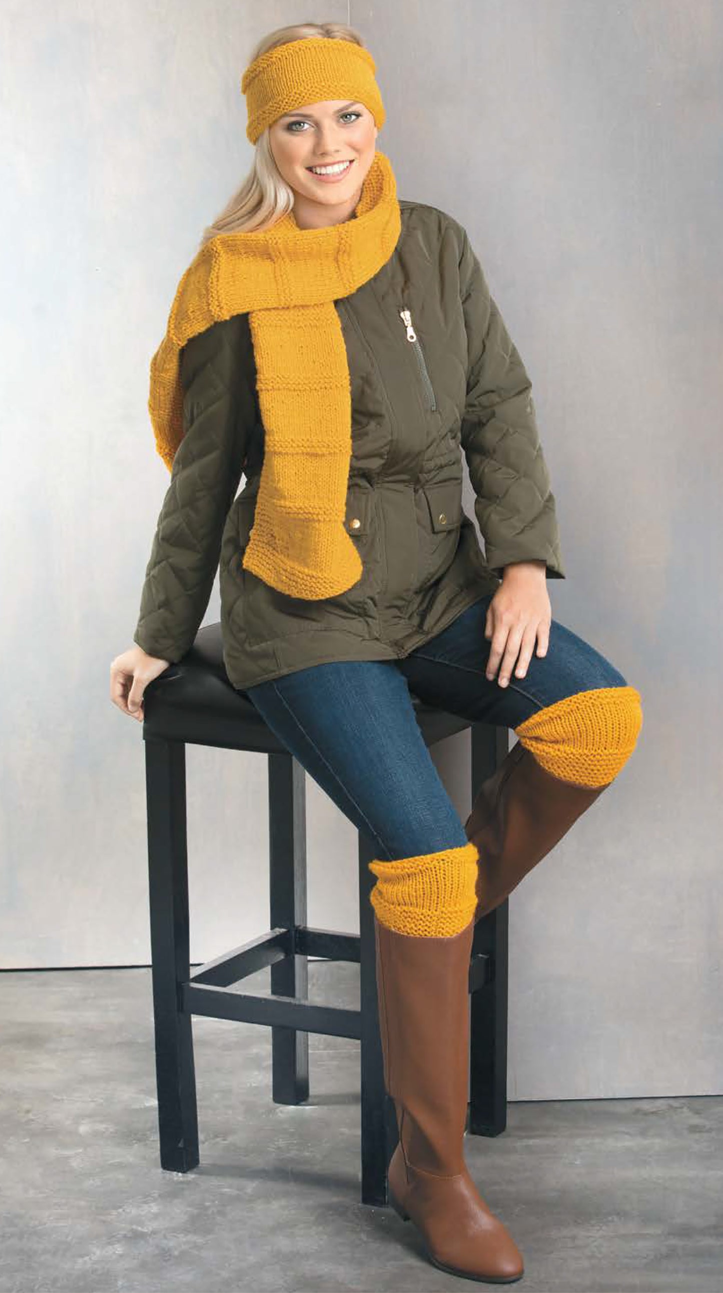 A woman models Cozy Fashion Accents from Leisure Arts, wearing mustard-yellow knit hat, scarf, and knee warmers. Styled with a green jacket and boots, she sits on a black stool, smiling at the camera. Digital download available.