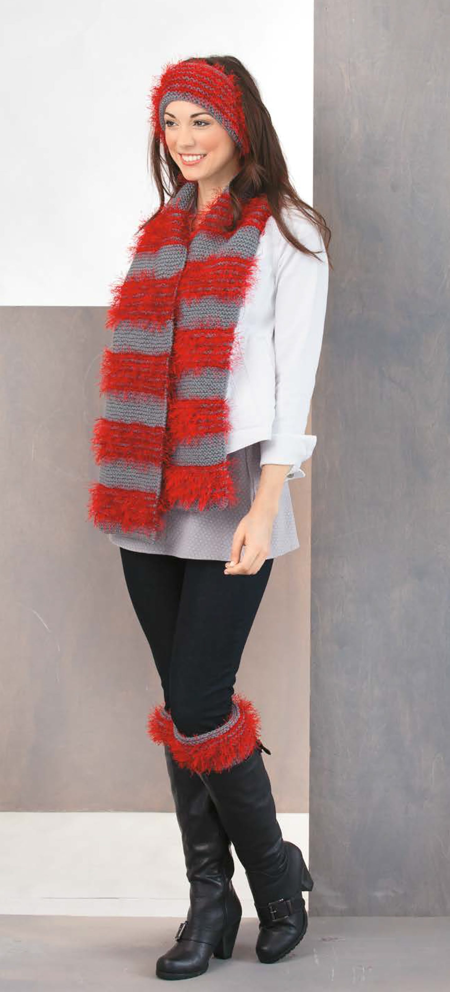 A woman smiles wearing a white shirt, black pants, and boots styled with Cozy Fashion Accents Digital Download by Leisure Arts—featuring a matching red and gray scarf, ear warmers, and fuzzy boot cuffs.