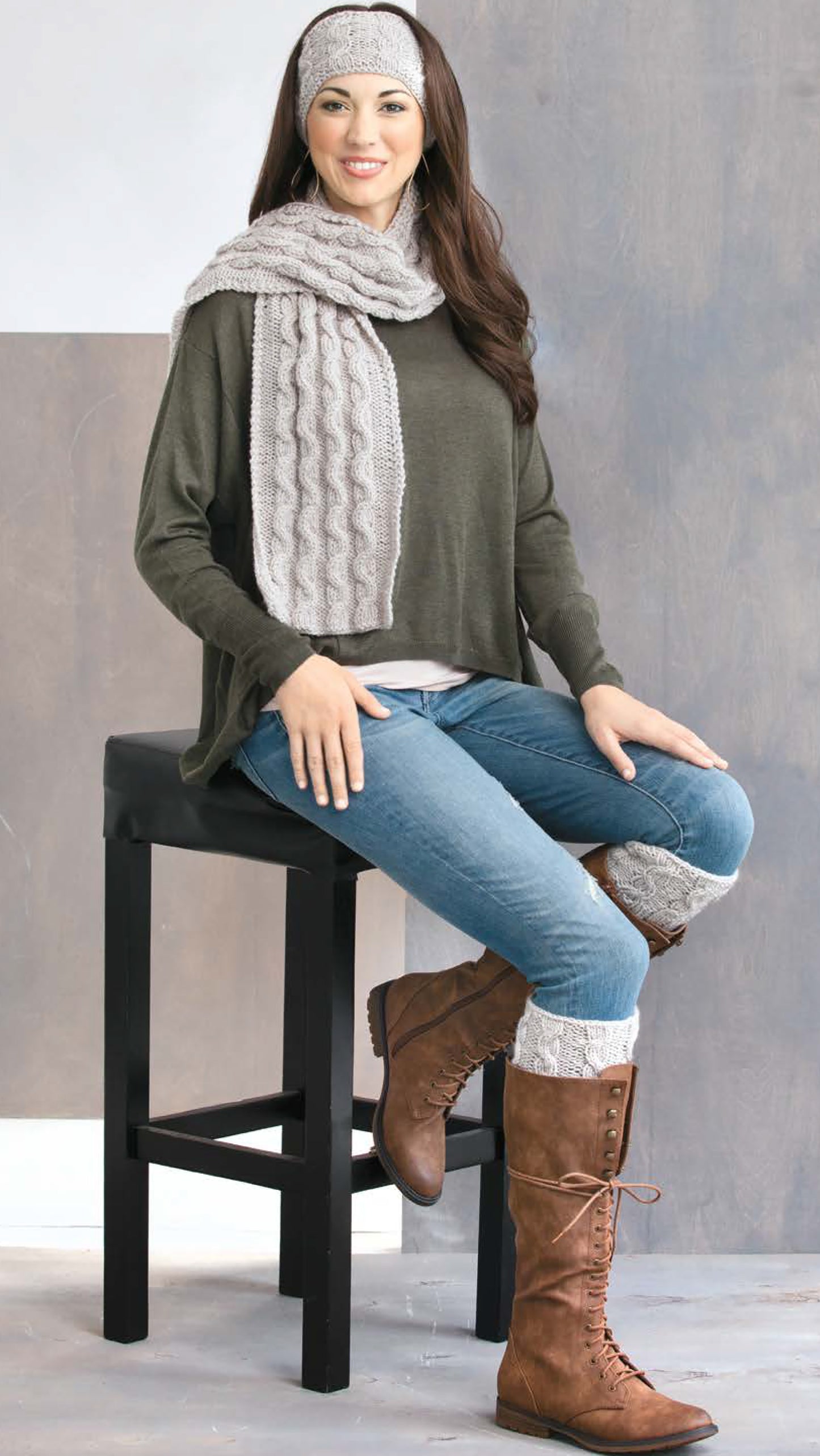 A woman models Cozy Fashion Accents by Leisure Arts: gray knit boot cuffs and ear warmers, styled with a green long-sleeve top, blue jeans, and brown lace-up boots while smiling on a black stool against a neutral background.