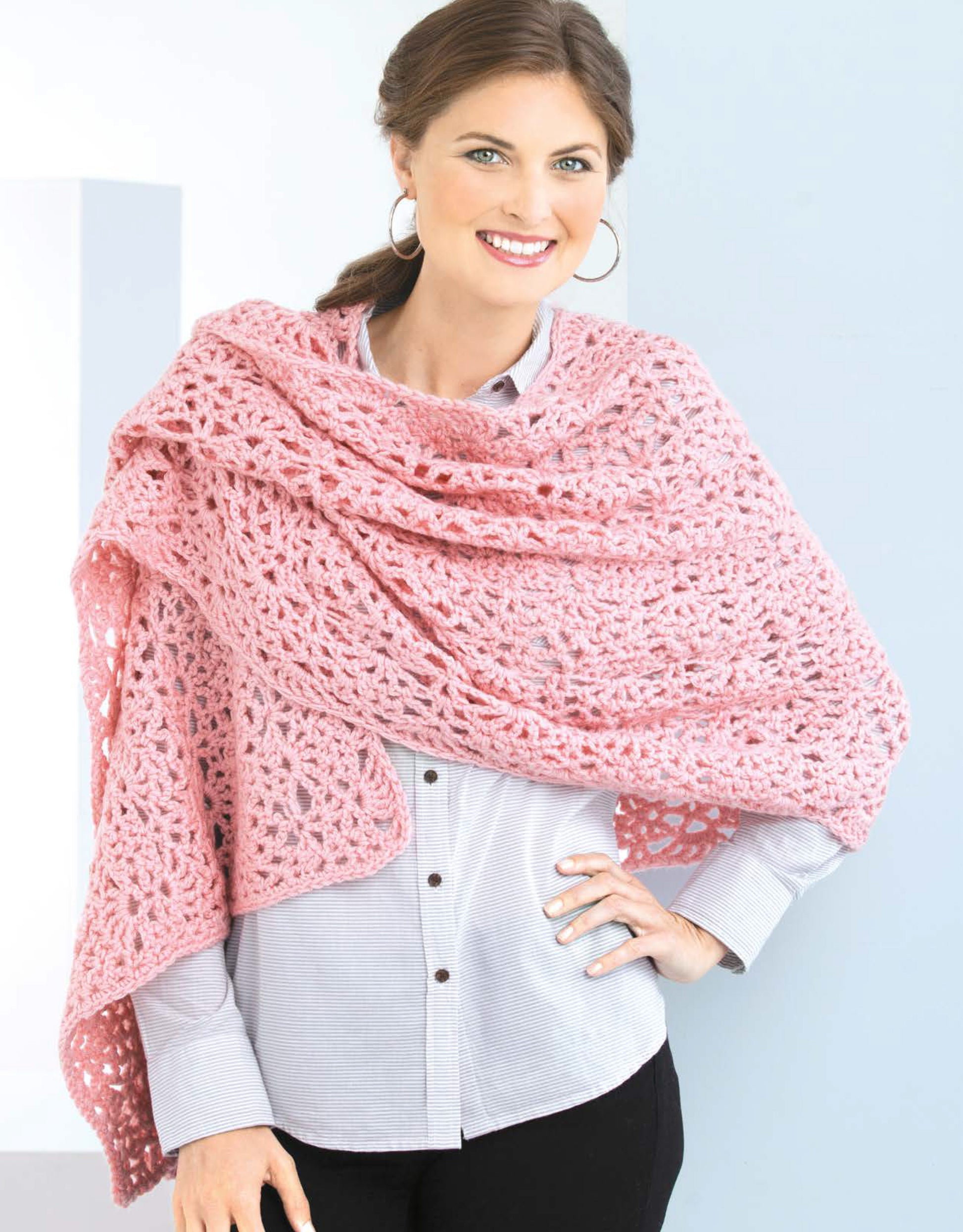 A smiling woman with brown hair wears a light blue button-up and a large pink crochet shawl, as featured in "Crochet Shawls You’ll Love" by Leisure Arts—perfect for those seeking stylish worsted weight crochet patterns.