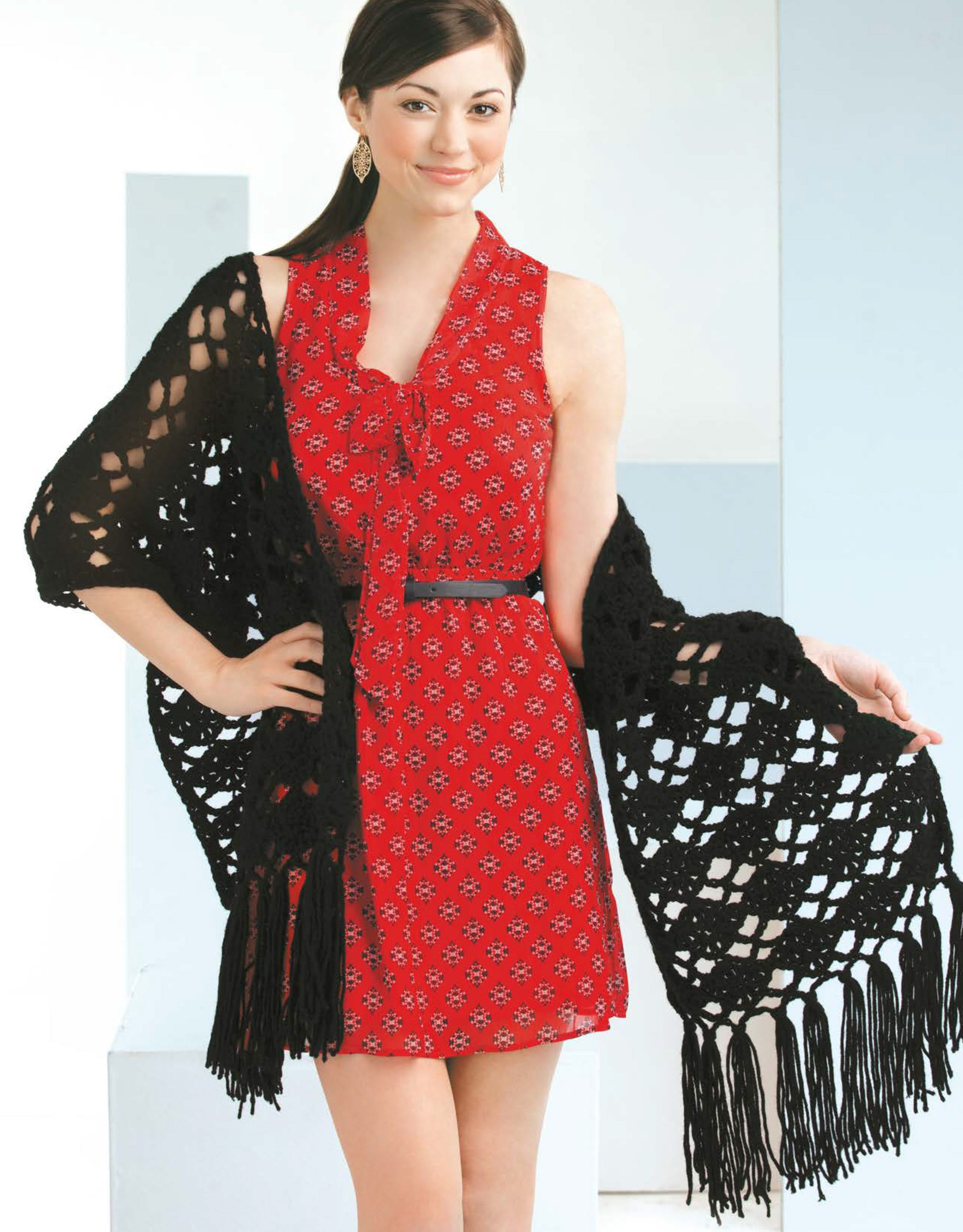 A woman models a black fringed shawl made from worsted yarn, featured in "Crochet Shawls You'll Love" by Leisure Arts, over a sleeveless red patterned dress while smiling at the camera against a light background.