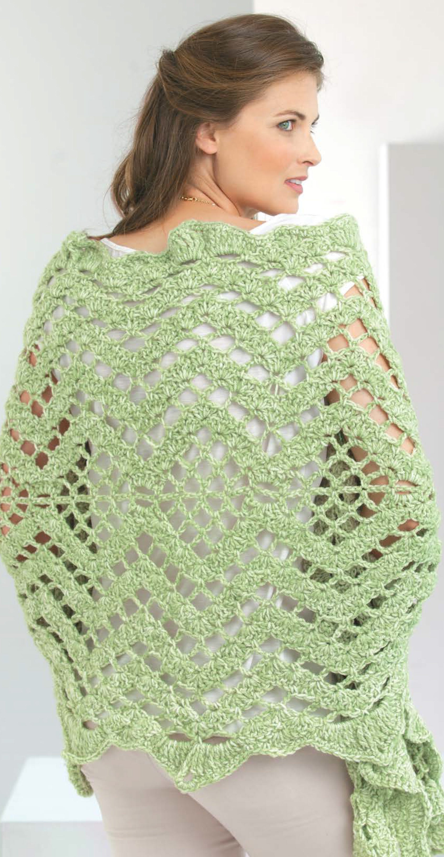 A woman with long brown hair models a large, light green crochet shawl with zigzag and openwork patterns from "Crochet Shawls You'll Love" by Leisure Arts, worn over a white top and light pants in a bright, neutral room.