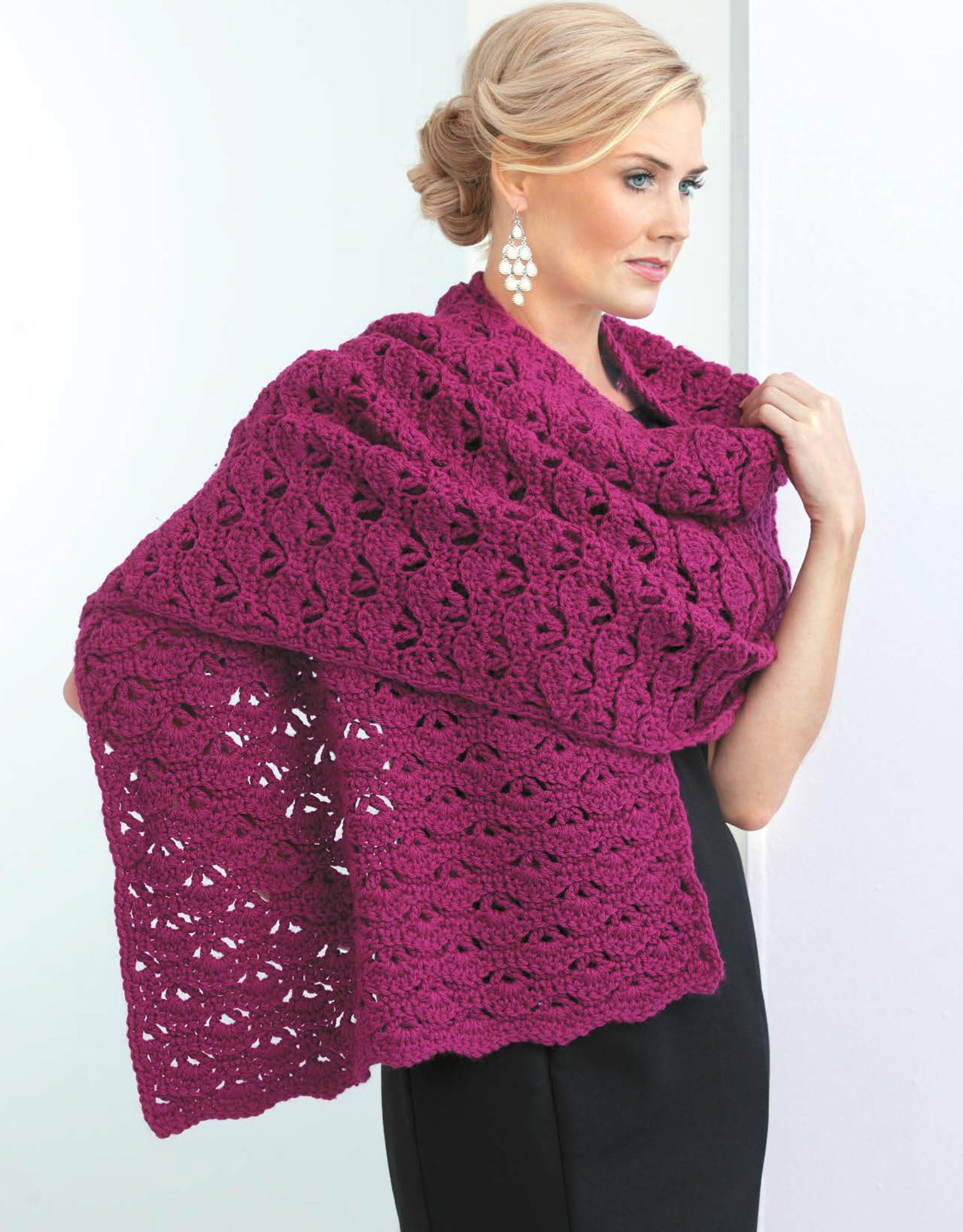 A woman in a sleeveless black dress models a large, textured deep magenta crochet shawl from "Crochet Shawls You'll Love" by Leisure Arts, draped over her shoulders. She wears dangling earrings and her blonde hair is styled in an elegant updo.