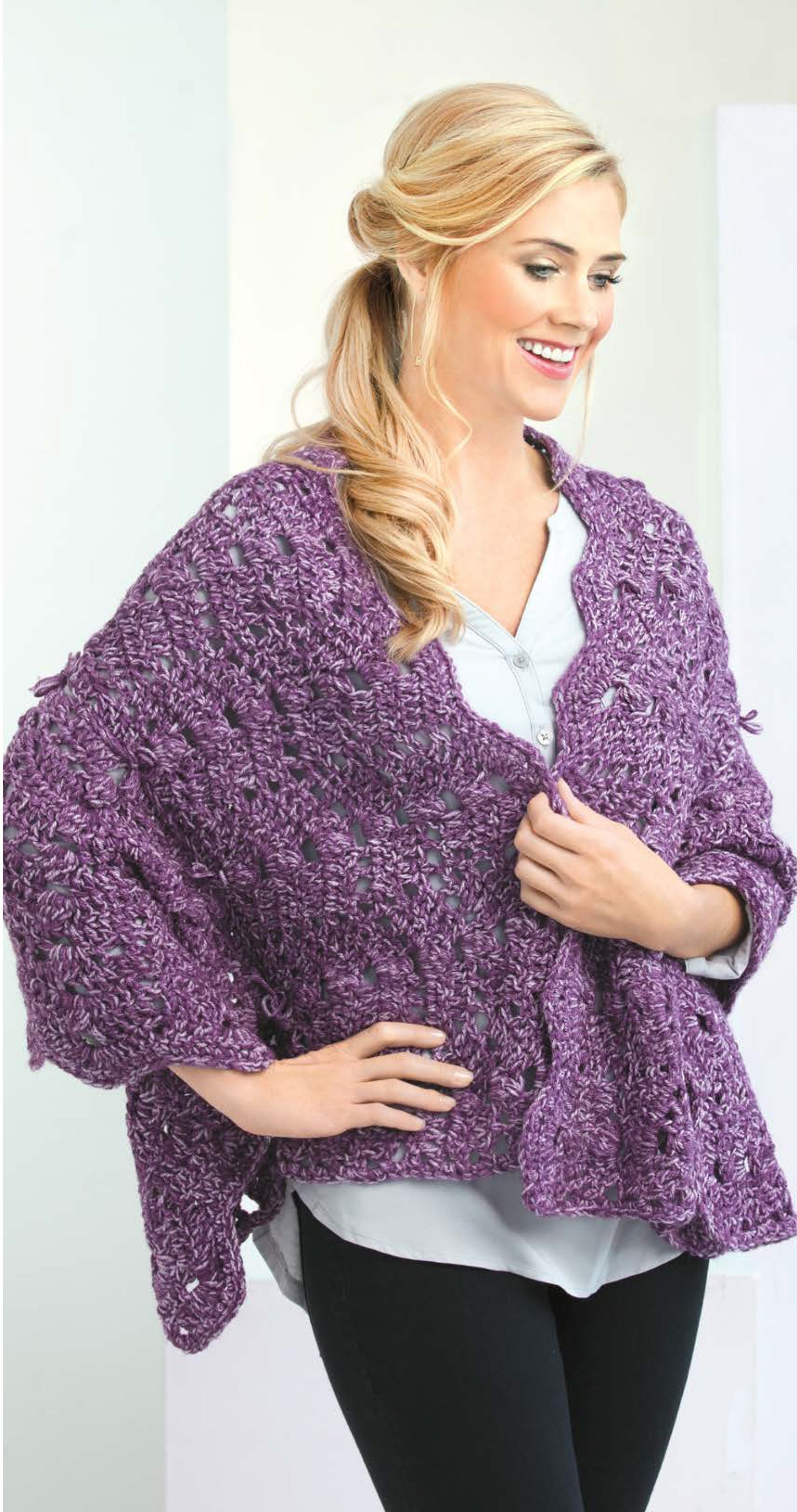 A smiling woman with long blonde waves models a loose purple shawl from Crochet Shawls You'll Love by Leisure Arts, draped over a light grey blouse and black pants, holding the front closed with one hand.