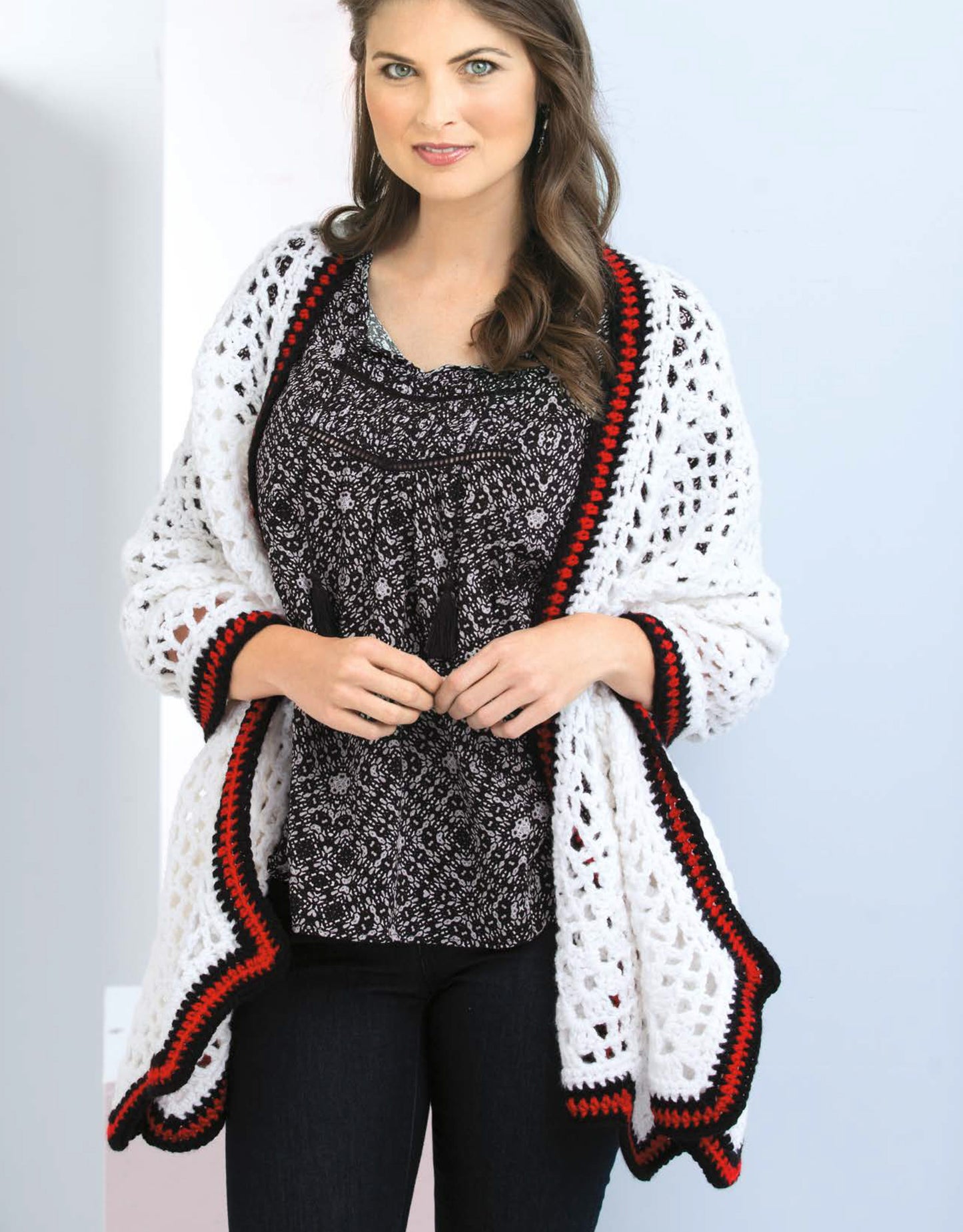 A woman with long brown hair models a white crocheted shawl with red and black trim from "Crochet Shawls You'll Love" by Leisure Arts, over a patterned blouse and black pants, against a light background.