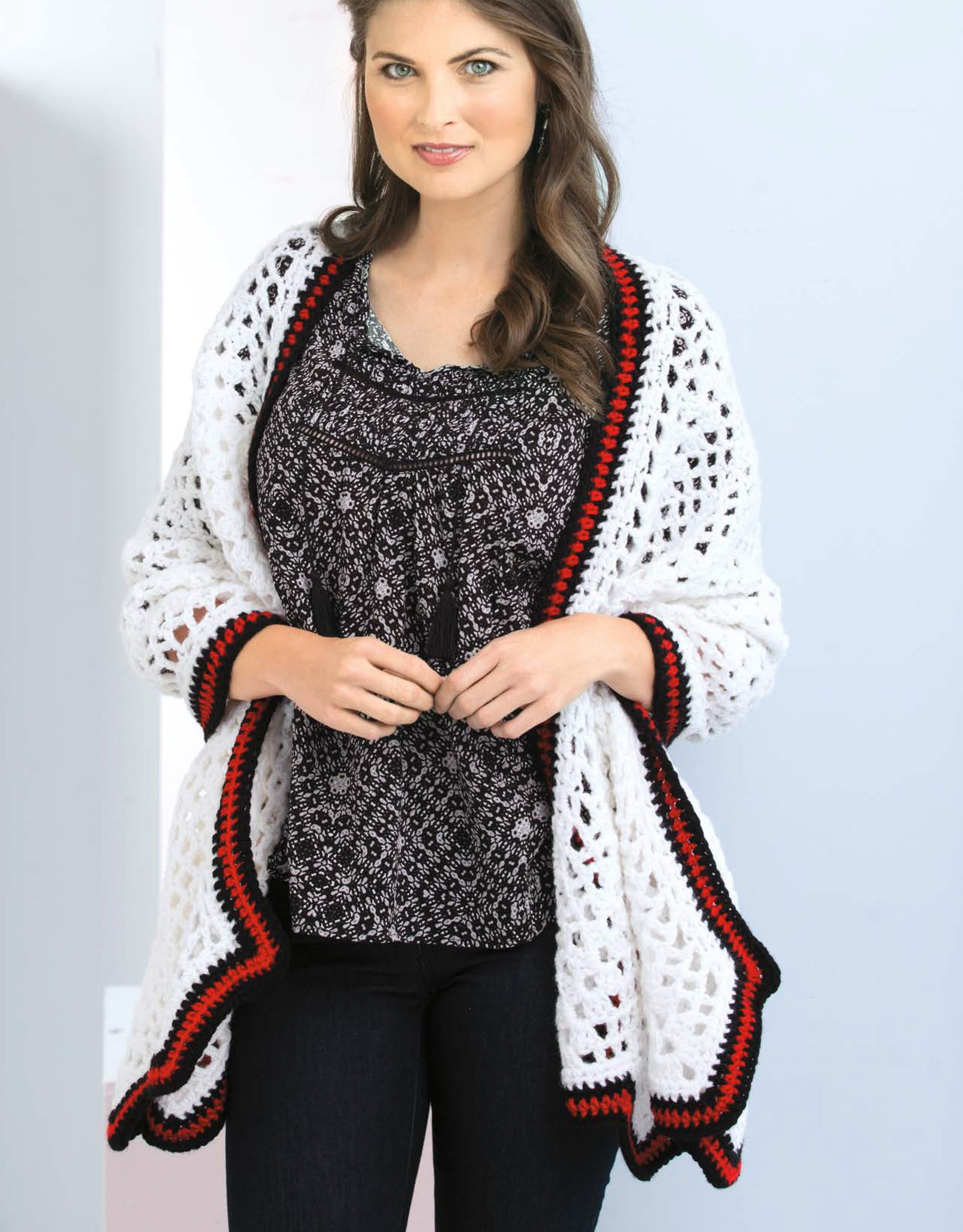 A woman with long brown hair models a white crocheted shawl with red and black trim from "Crochet Shawls You'll Love" by Leisure Arts, over a patterned blouse and black pants, against a light background.
