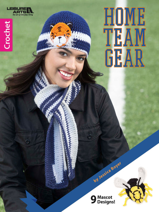 A smiling woman wears a blue and white striped crochet scarf and a tiger face hat. Text reads Home Team Gear by Leisure Arts—9 mascot designs! Show school spirit with cheerful crochet mascots, like a friendly bee.
