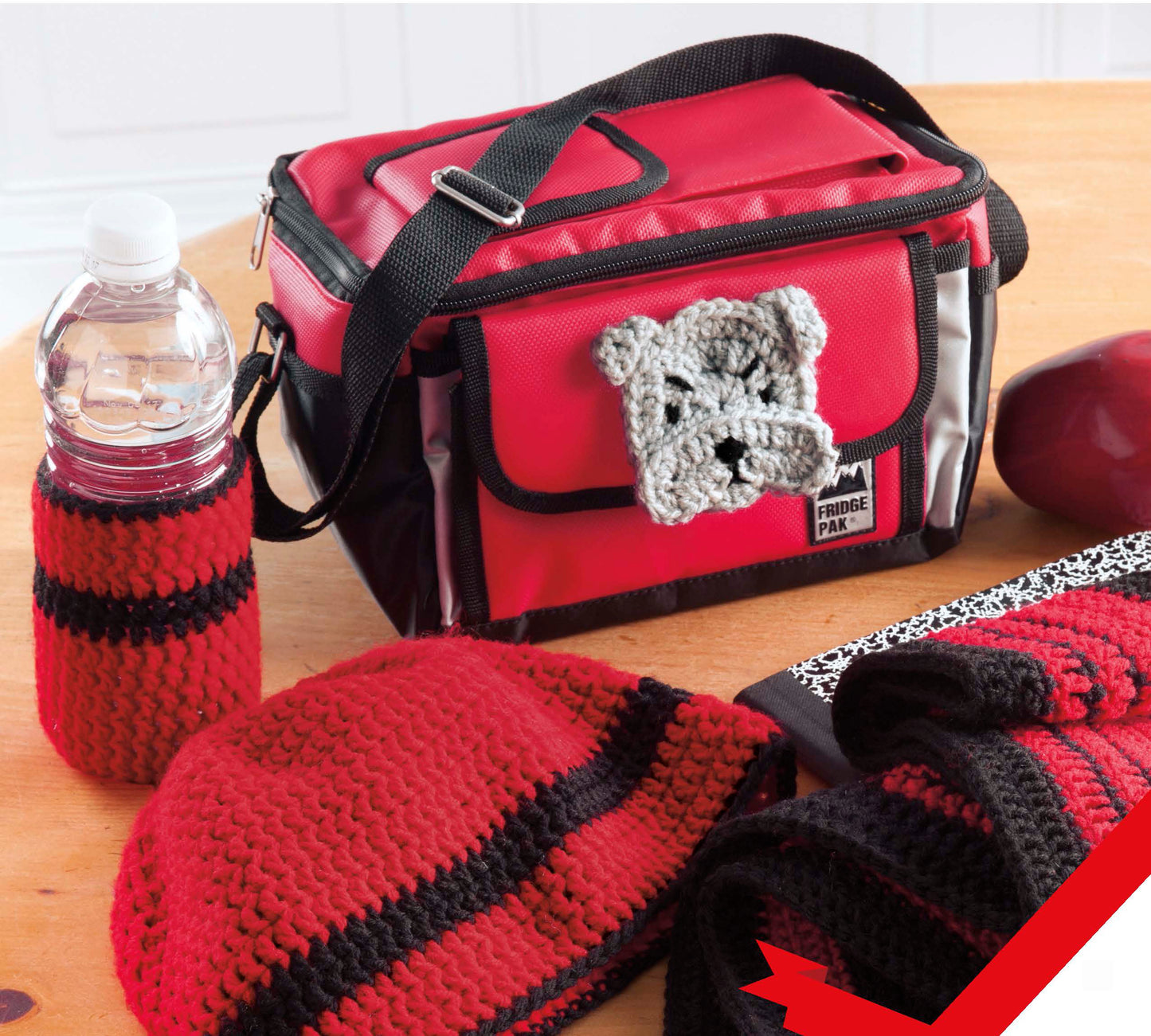 Home Team Gear by Leisure Arts—a red and black insulated lunch bag with a crocheted bulldog face, plus a matching water bottle holder, hat, and scarf—sits on the table, showing off perfect school spirit style.