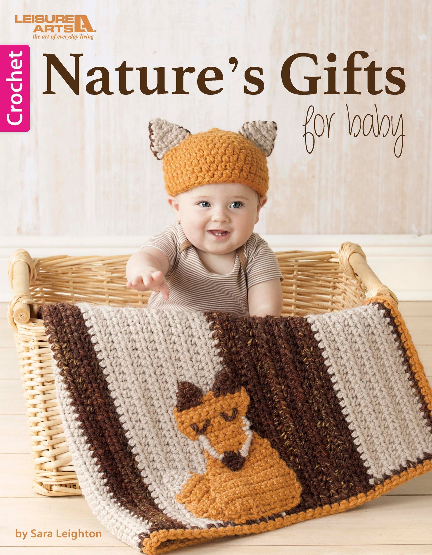 A smiling baby in a crocheted fox ear hat sits in a basket behind a Nature's Gifts for Baby quilt by Leisure Arts, featuring an adorable fox design.