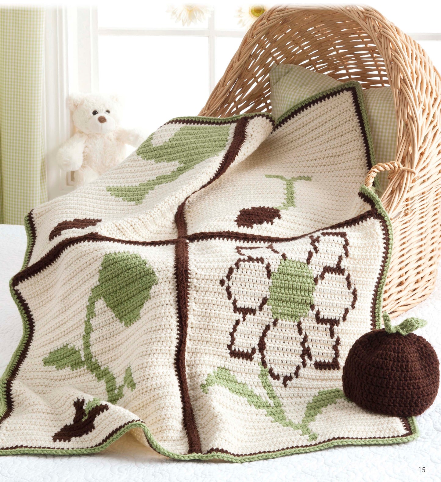 Nature's Gifts for Baby" by Leisure Arts is a cream knit blanket with green and brown nature designs, displayed draped over a wicker basket beside a crochet bulb and white teddy bear—ideal as a cozy, nature-inspired baby quilt.