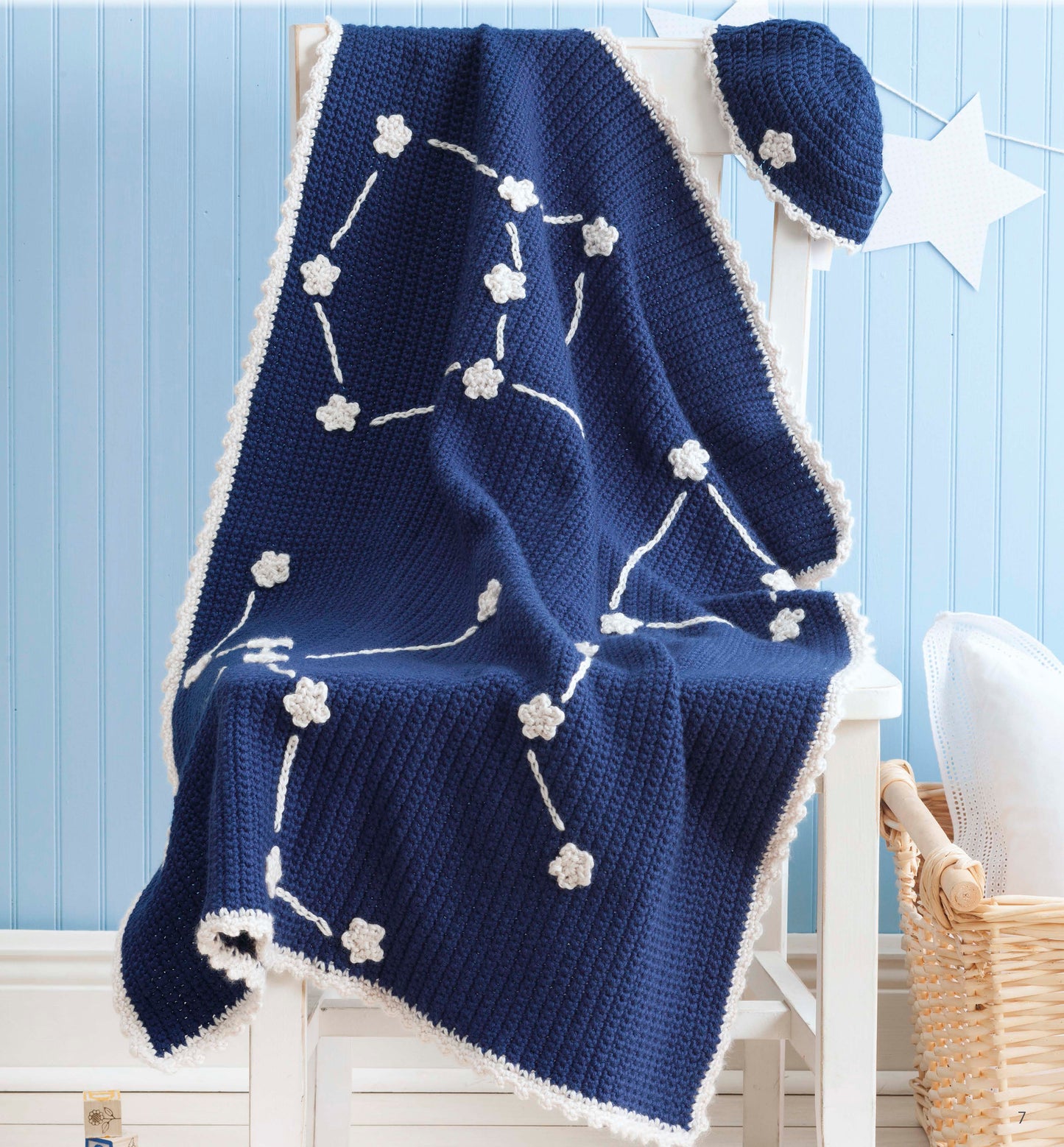 The Nature's Gifts for Baby set by Leisure Arts features a navy blue knitted baby blanket with water lilies, white stars, and constellations, draped over a white chair with a matching hat, all set against a light blue paneled wall.