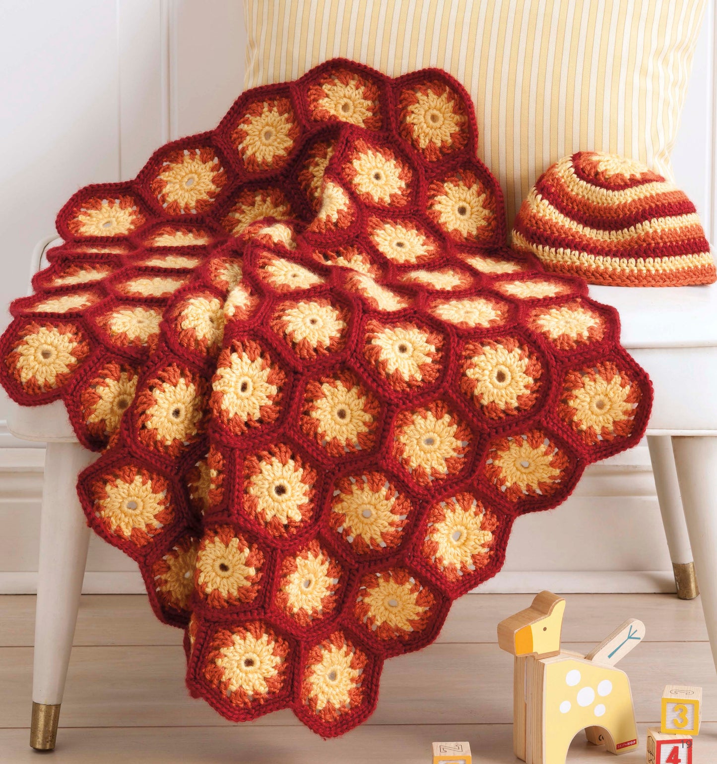 Nature's Gifts for Baby by Leisure Arts features a red and yellow hexagon blanket draped over a white chair, with a matching striped hat, pillow, wooden giraffe toy, and letter blocks placed on the floor.