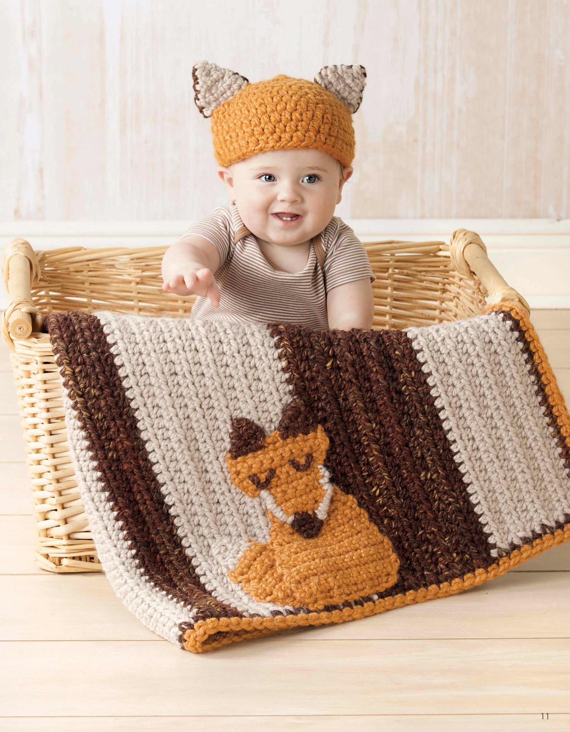A smiling baby in a crocheted orange fox-ear hat sits in a wicker basket, draped with the Nature's Gifts for Baby quilt by Leisure Arts, adding cozy woodland charm to the scene.