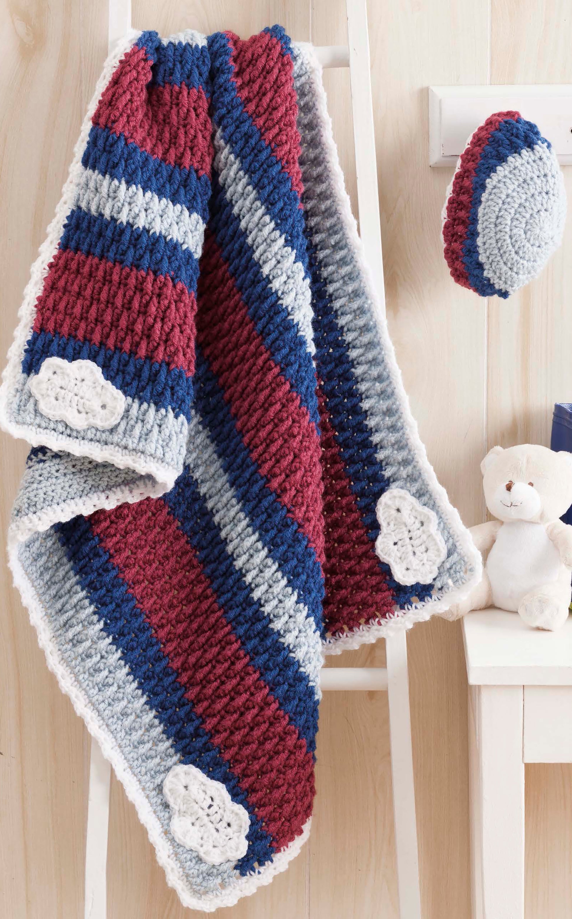 Nature's Gifts for Baby by Leisure Arts features a crocheted water lilies baby blanket with red, blue, and gray stripes plus white cloud appliqués, draped over a white ladder with a matching hat and teddy bear against a light wood background.