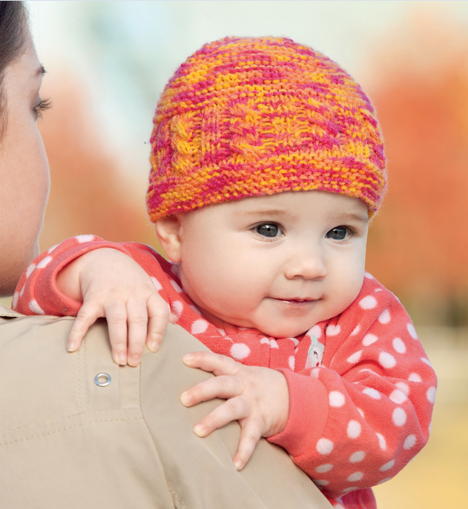 A baby in a pink polka-dot outfit wears a colorful hat made with the "Hats and Scarves for Kids" digital download by Leisure Arts, held by an adult outdoors amid softly blurred autumn trees.