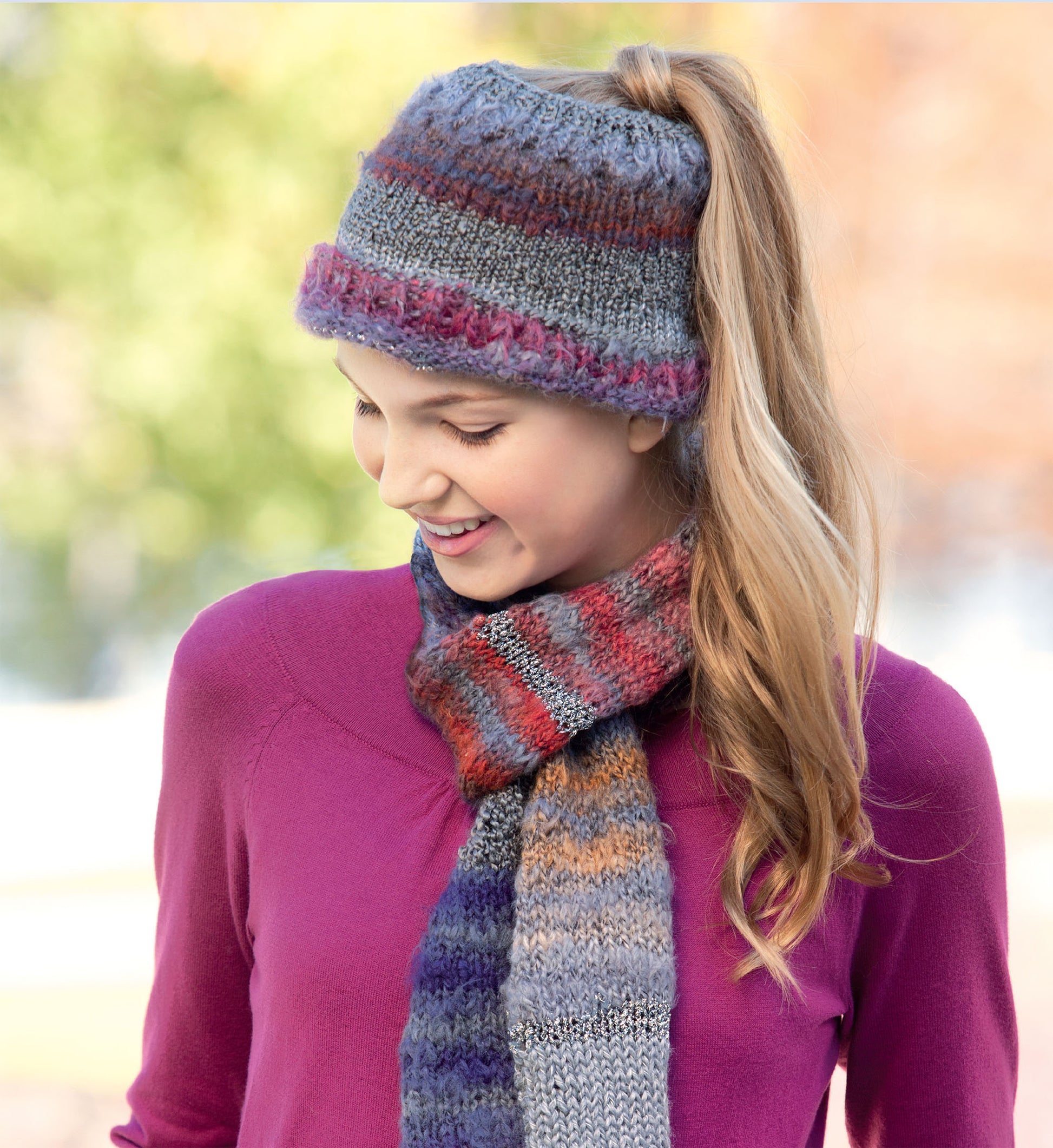 A young woman in a purple sweater and knit hat with a ponytail opening smiles while wearing a striped scarf—perfect inspiration for easy projects from Leisure Arts’ "Hats and Scarves for Kids" digital download. Blurred outdoor greenery is in the background.