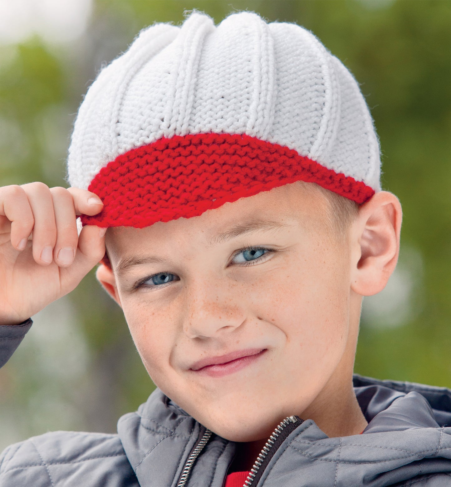 A boy outdoors in a quilted jacket smiles as he adjusts his red and white knitted cap—an ideal project from the "Hats and Scarves for Kids Digital Download" by Leisure Arts, with green trees softly blurred in the background.