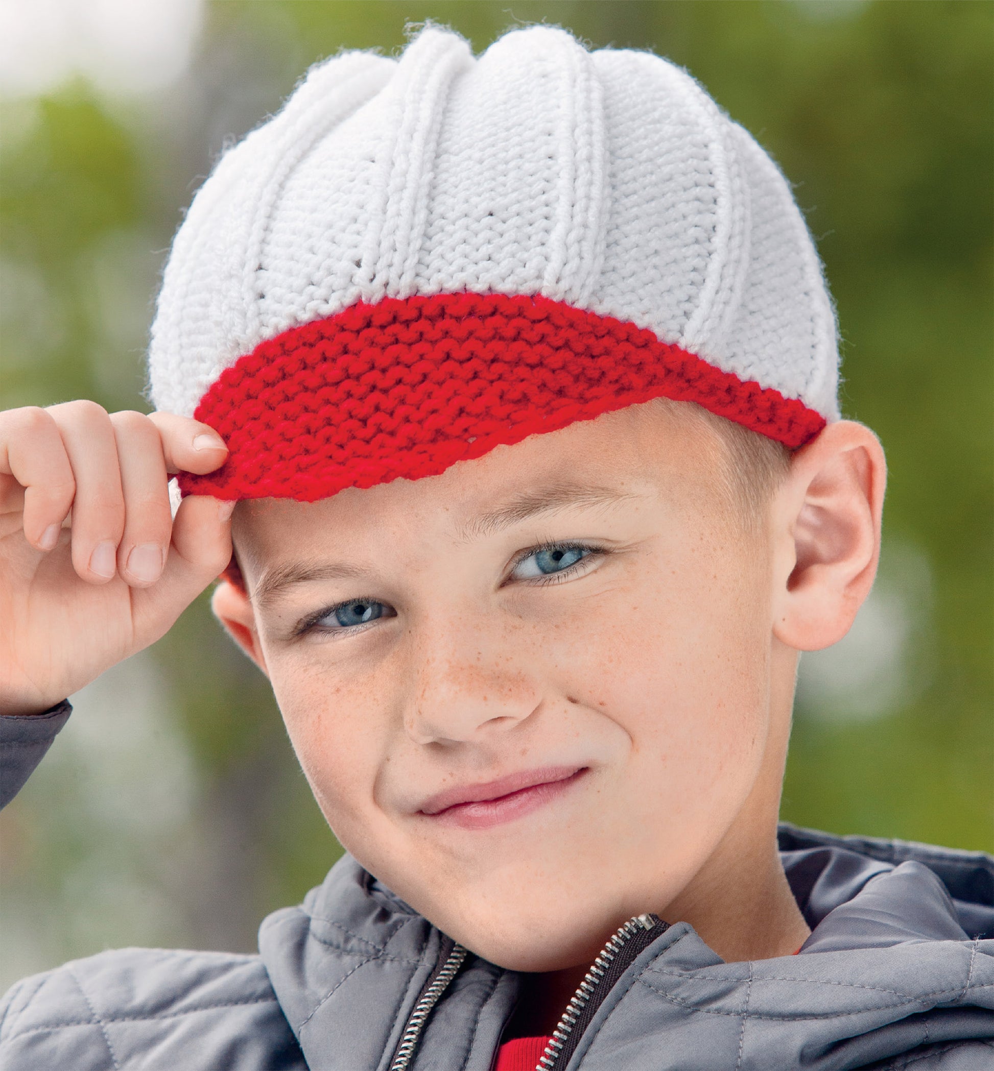 A boy outdoors in a quilted jacket smiles as he adjusts his red and white knitted cap—an ideal project from the "Hats and Scarves for Kids Digital Download" by Leisure Arts, with green trees softly blurred in the background.